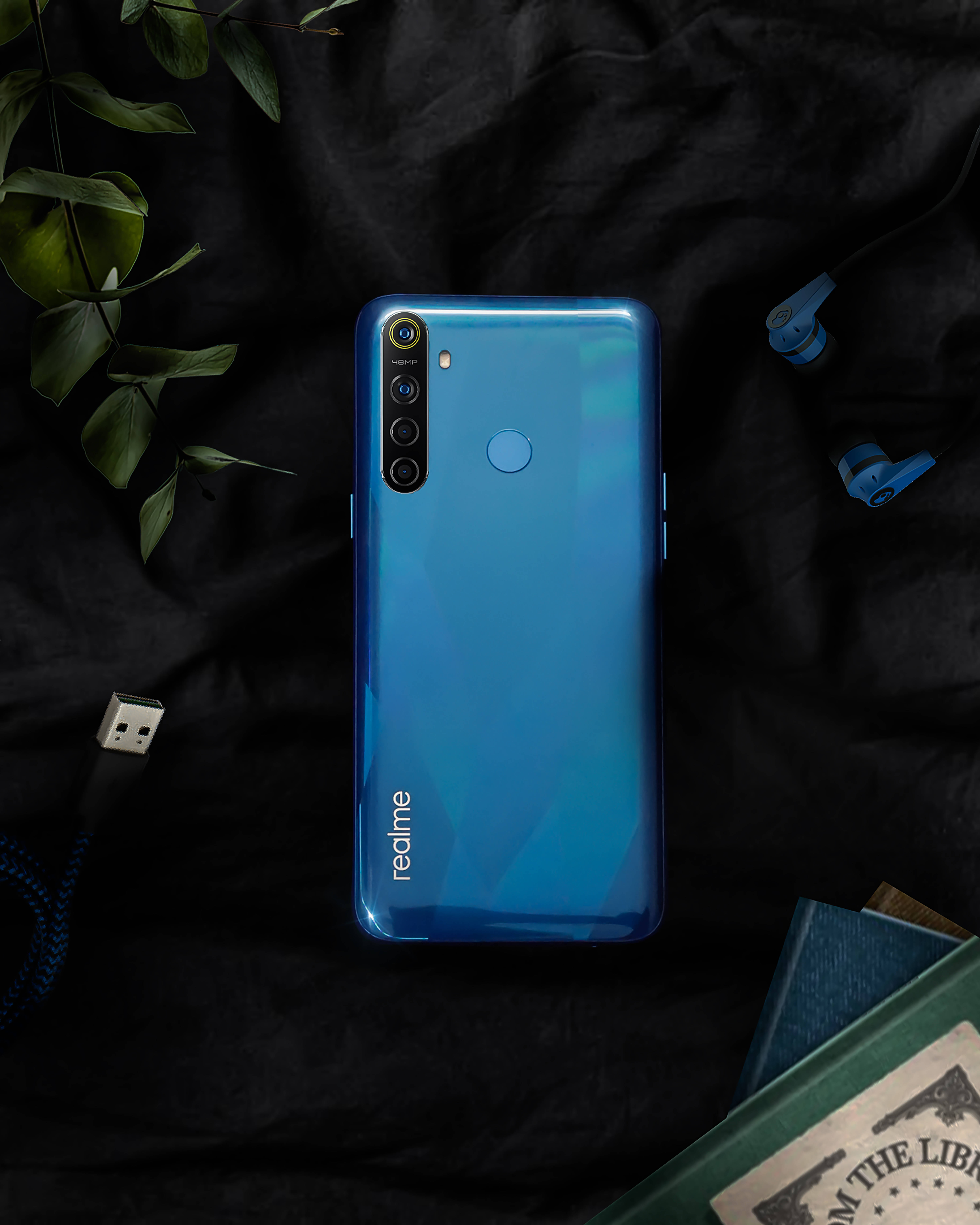 Realme 8 手機 壁紙 — 一部亮藍色 realme 智慧型手機放在黑色布料上，周圍有綠色樹葉、耳塞和一本書。