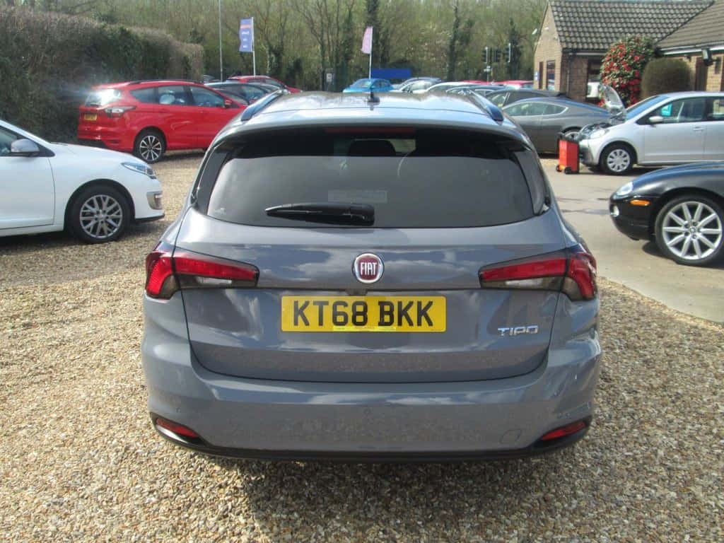 Fiat Tipo 桌布 壁紙 — 從後方觀看一輛灰色 Fiat Tipo 旅行車停放在陽光普照的停車場碎石地上，附近有其他車輛。