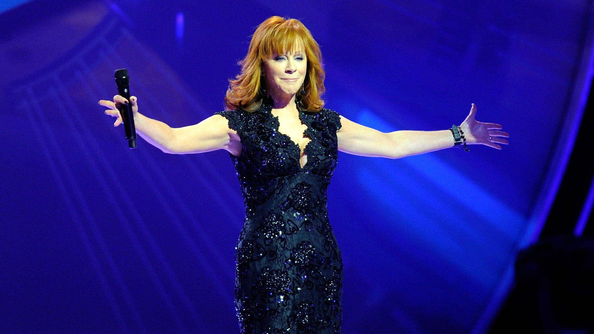 Celebrity 桌布 壁紙 — Reba McEntire 在舞台上穿著閃亮的黑色連衣裙，雙臂張開，在戲劇性的藍色舞台燈光下表演。