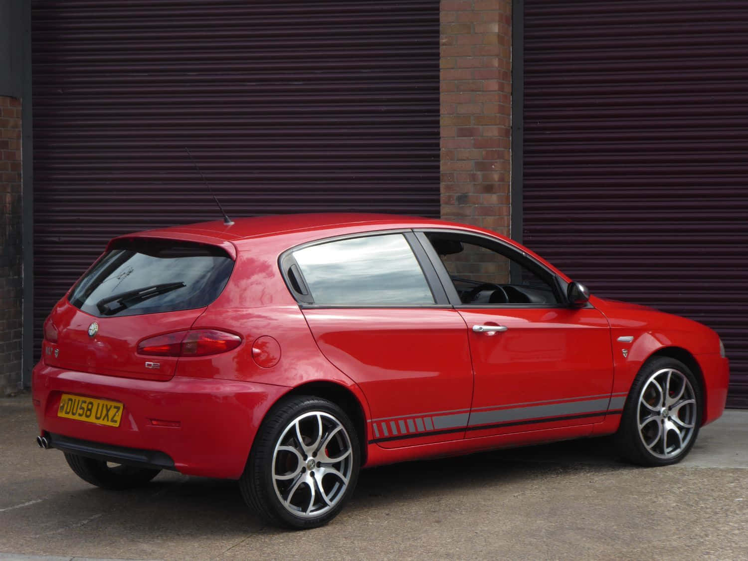 Alfa Romeo 147 桌布 壁紙 — 一輛鮮紅色的愛快羅密歐147掀背車，斜停在深紫色波紋金屬車庫門前。
