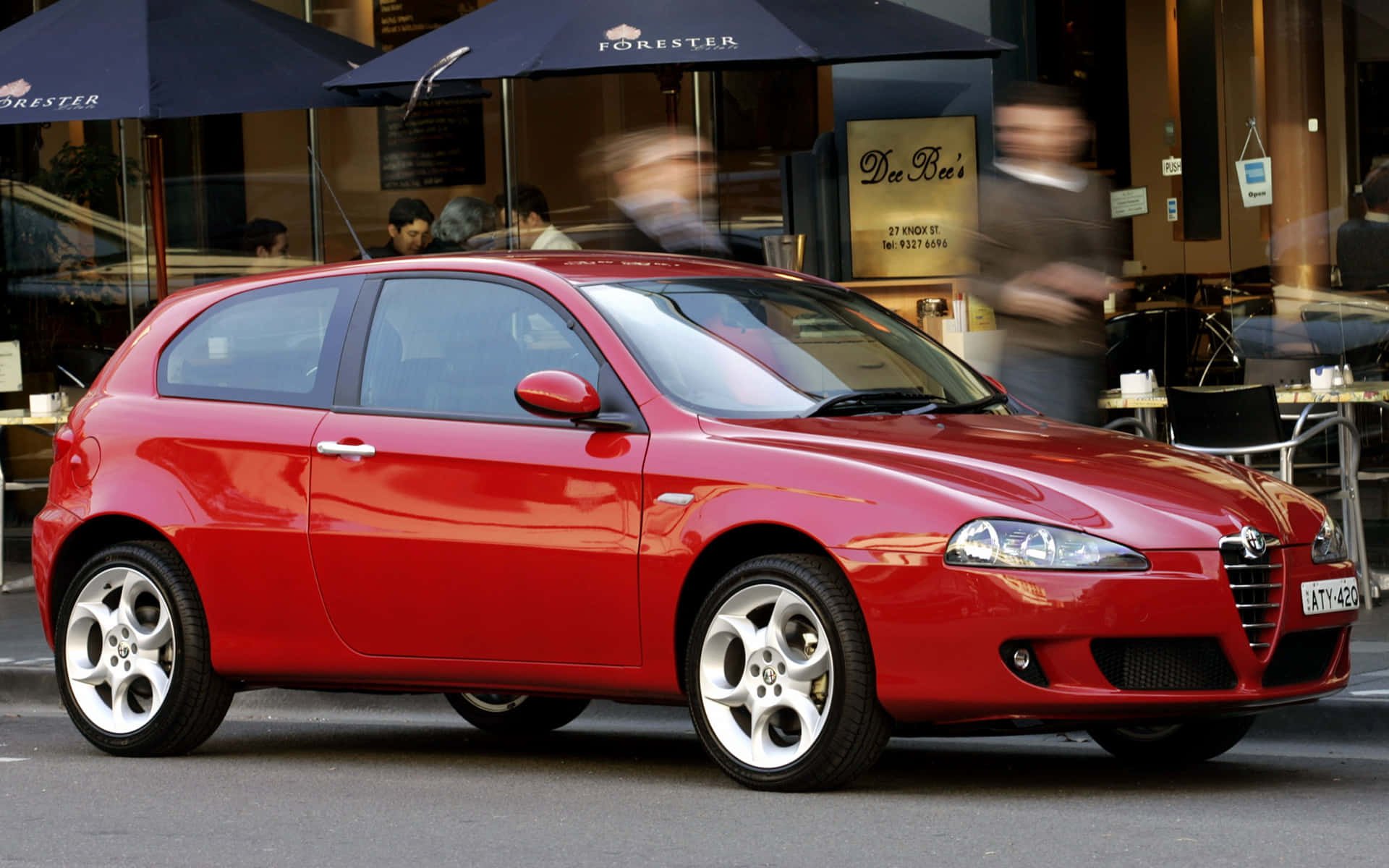Alfa Romeo 147 桌布 壁紙 — 一輛充滿活力的紅色愛快羅密歐147掀背車停泊在繁忙的城市街道上，背景有模糊的行人和咖啡館的遮陽傘。