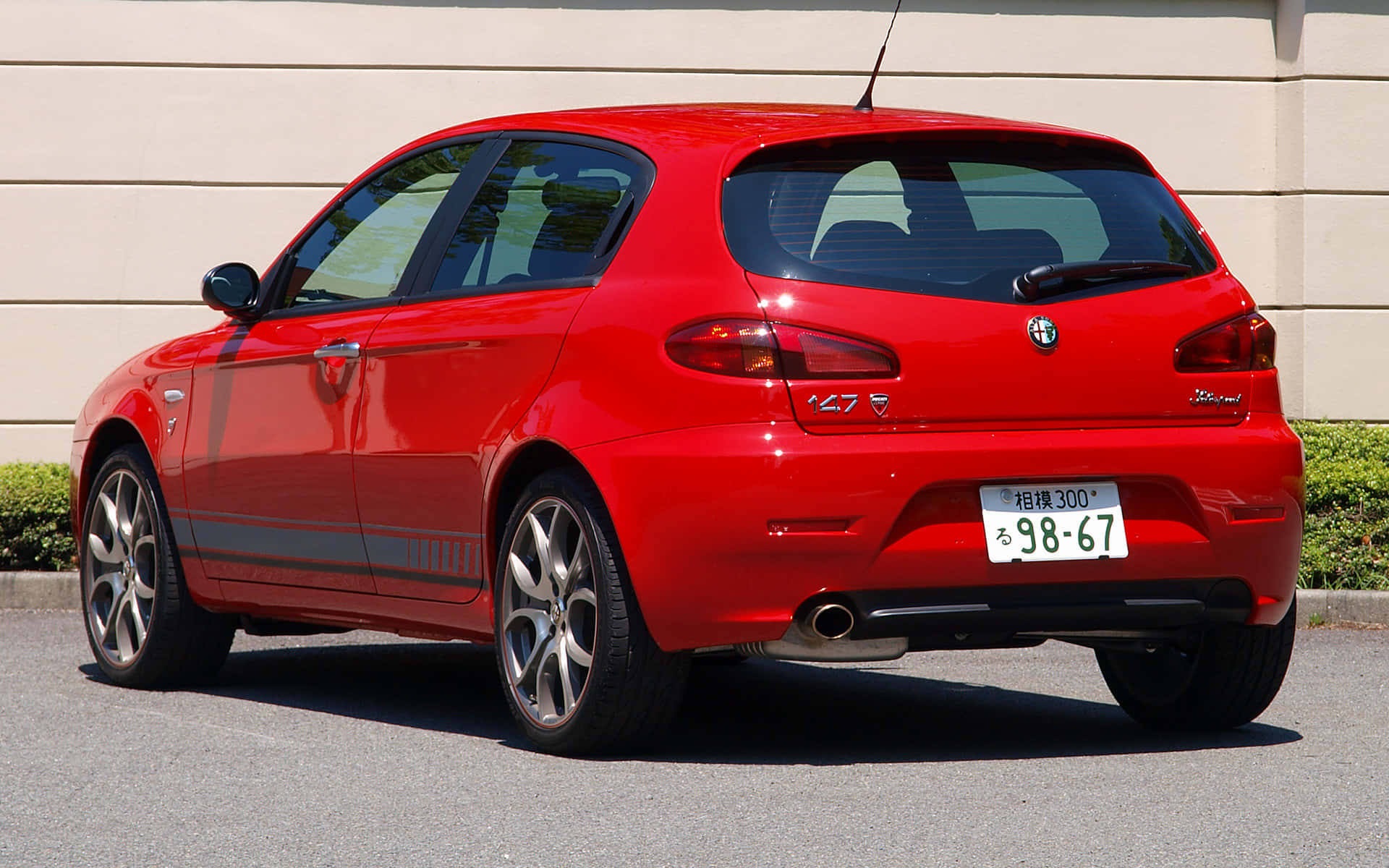 Alfa Romeo 147 桌布 壁紙 — 一輛鮮紅色的愛快羅密歐147跑車在陽光明媚的日子停放在戶外，背景有米色牆壁和綠色灌木。