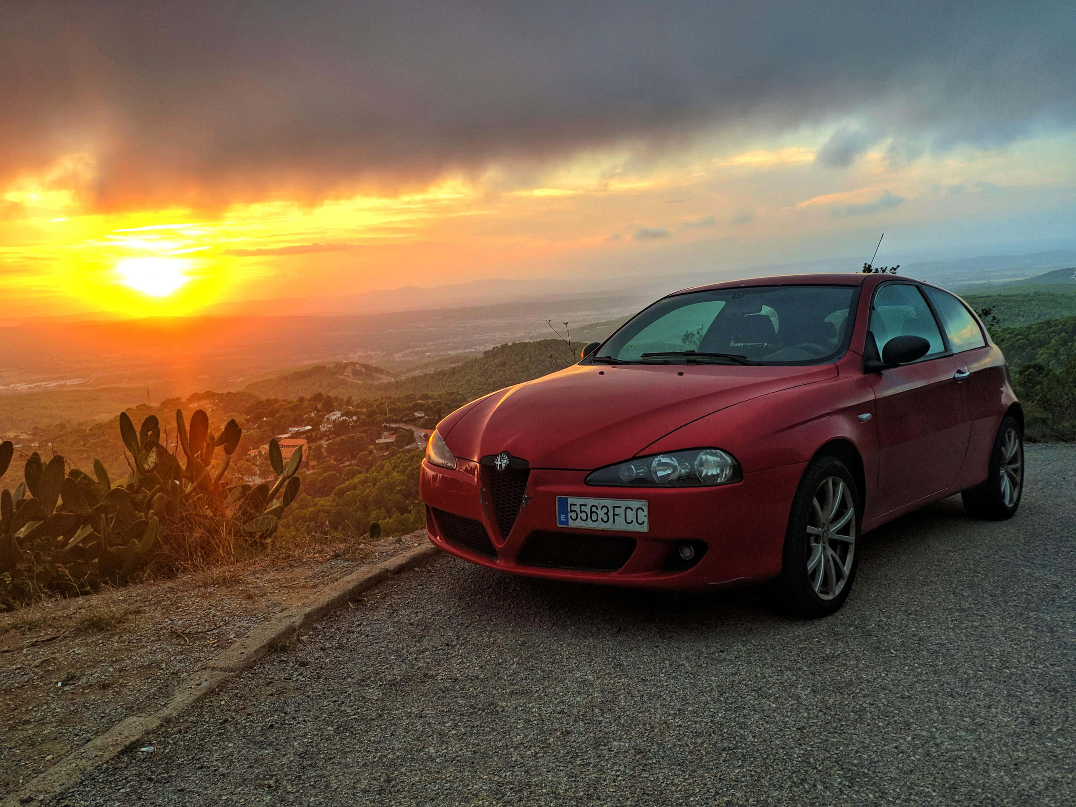 Alfa Romeo 147 桌布 壁紙 — 一輛紅色愛快羅密歐停泊在山坡路上，夕陽在遠山後落下，灑下溫暖的金色光芒。