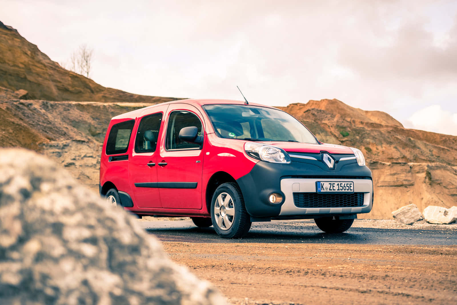 Renault Kangoo 桌布 壁紙 — 一輛紅色和黑色的雷諾Kangoo廂型車停放在碎石路上，背景是岩石丘陵，天空多雲。