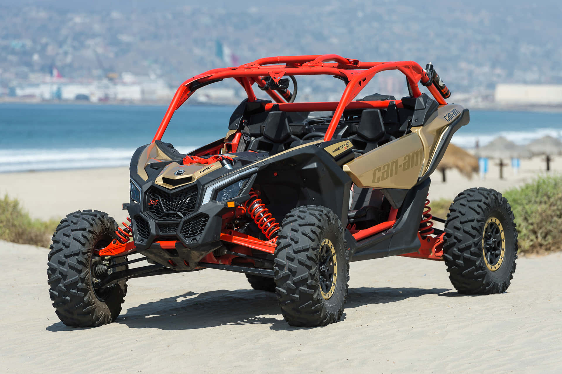  壁紙 — 一輛紅色和金色的Can-Am Maverick X3並排UTV停泊在沙灘上，背景是海洋和城市景觀。