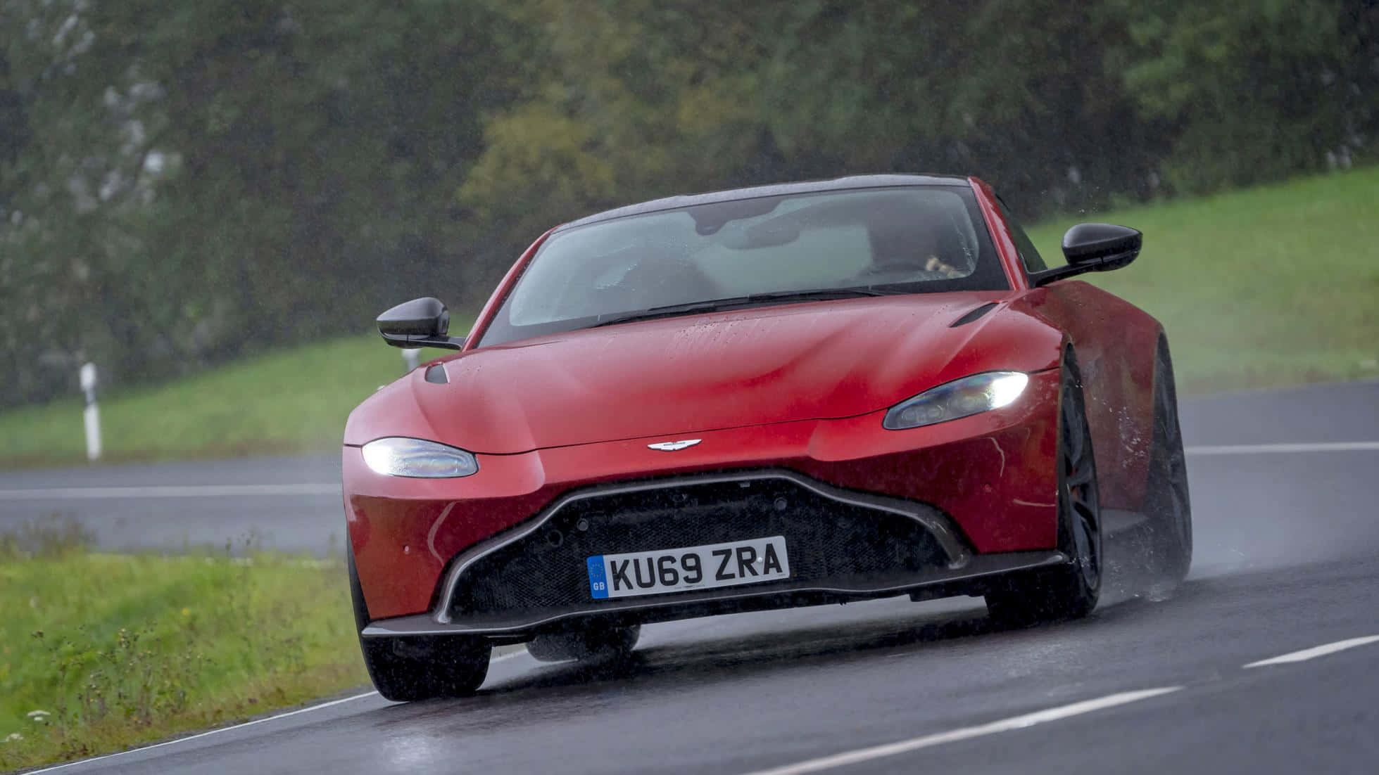 Aston Martin Vantage 桌布 壁紙 — 一輛紅色Aston Martin跑車在雨天沿著濕滑的道路穿過綠色鄉村飛馳。