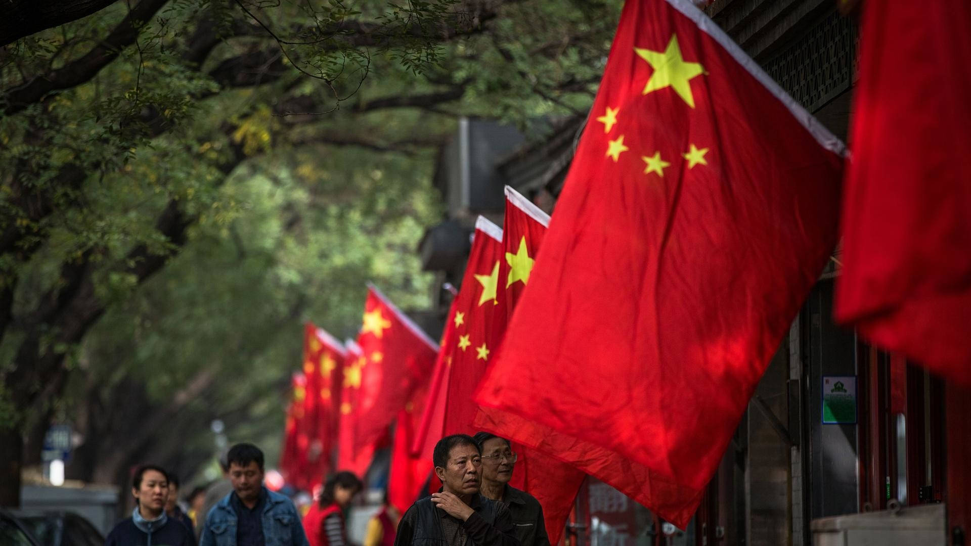 紅色中國國旗懸掛在一條綠樹成蔭的城市街道上，行人經過。 — 中國國旗 壁紙