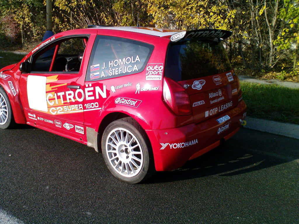 Citroen C2 Super 1600 桌布 壁紙 — 一輛鮮紅色的雪鐵龍C2 Super 1600拉力賽車停在瀝青路上，背後是秋日樹木在金色陽光下閃耀。
