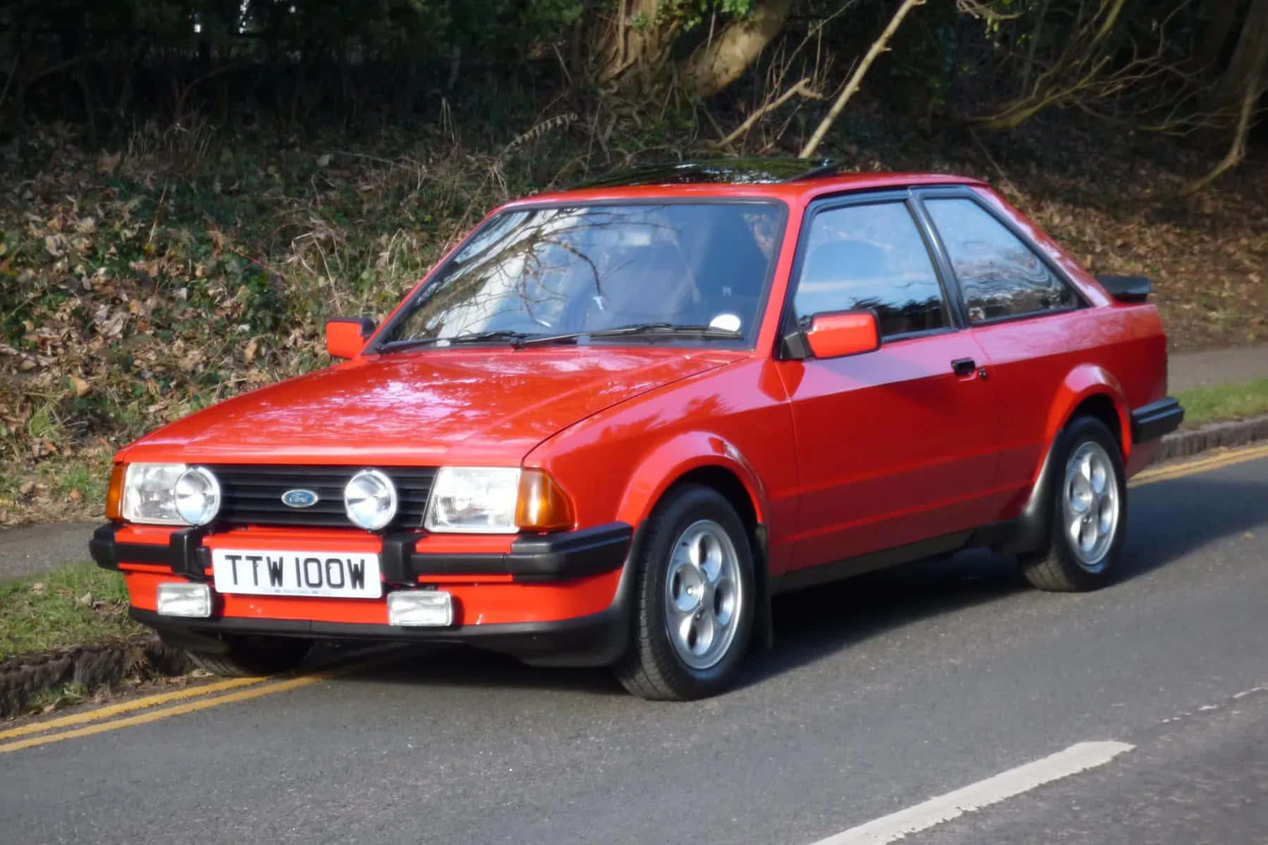 Ford Escort 桌布 壁紙 — 一輛鮮紅色的經典福特 Sierra Cosworth 停放在路邊，背景是沒有葉子的樹木和草地。