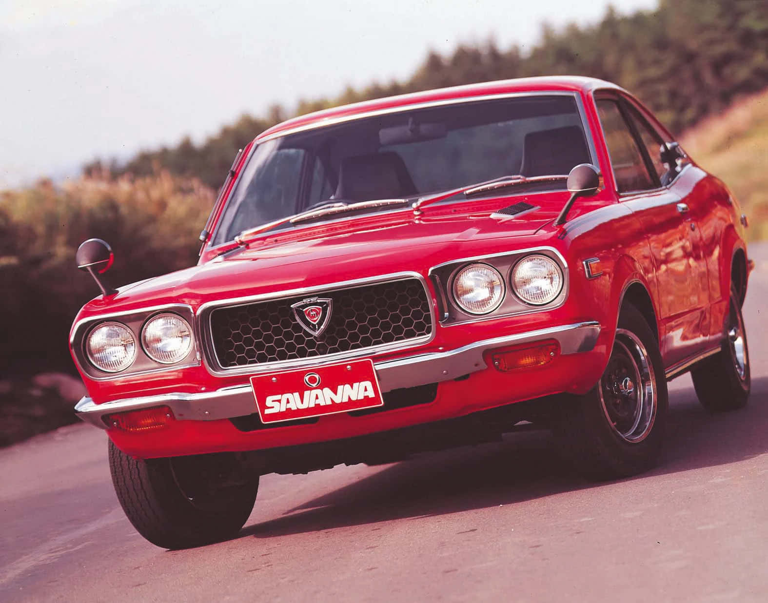 Mazda Rx-3 桌布 壁紙 — 一輛鮮紅色的經典Savanna轎跑車在明亮的陽光下閃閃發光，沿著蜿蜒的鄉村道路行駛。
