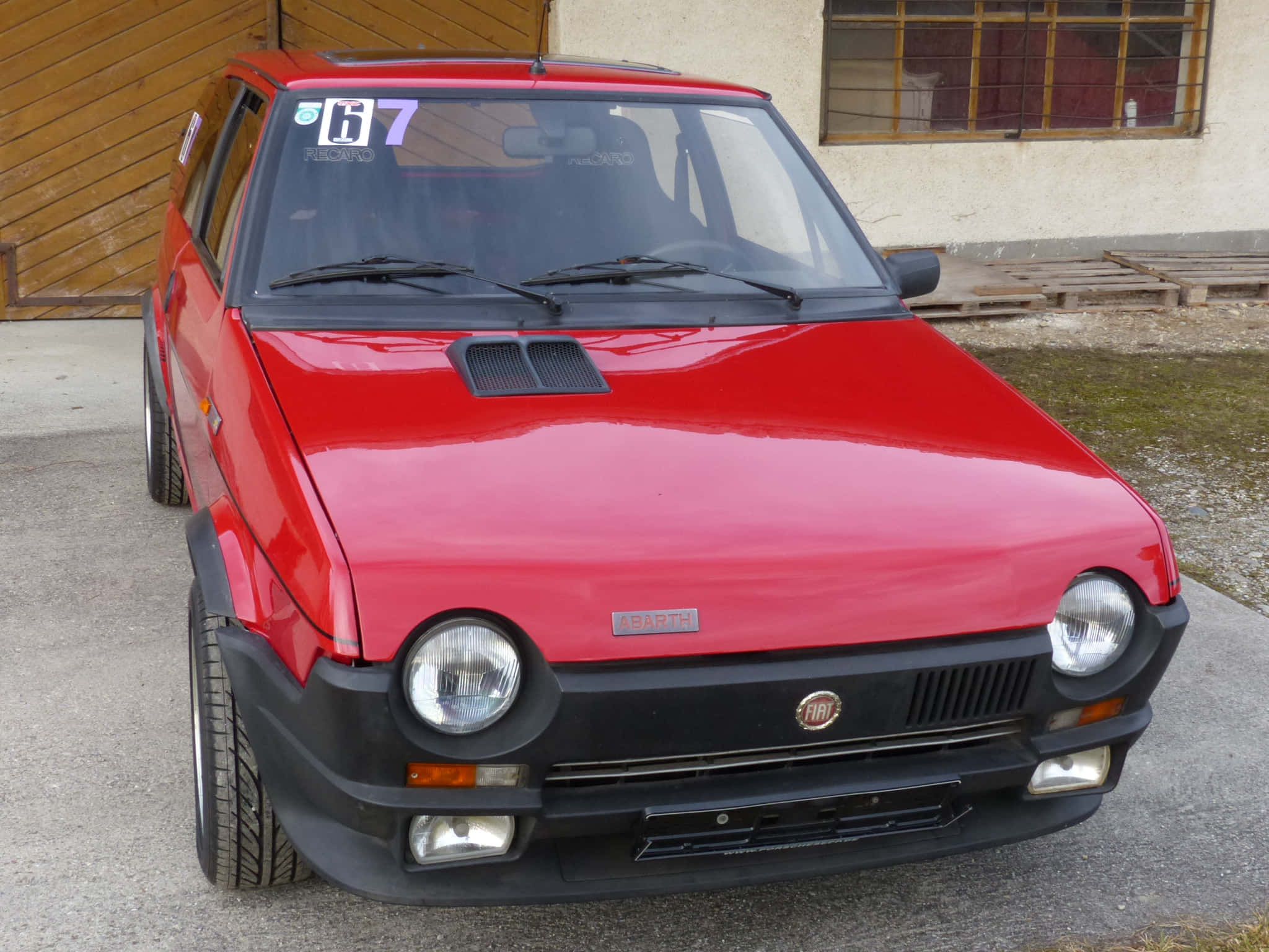 Fiat Ritmo 桌布 壁紙 — 一輛鮮紅色的Fiat 126p拉力賽車，帶有賽車號碼67，停在木車庫前的混凝土上。