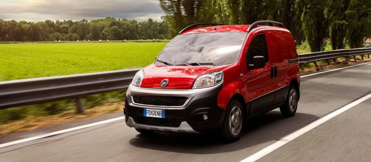 Fiat Fiorino 桌布 壁紙 — 一輛鮮紅色的菲亞特貨車在陰天下一條兩旁長滿翠綠田野和樹木的高速公路上疾馳。