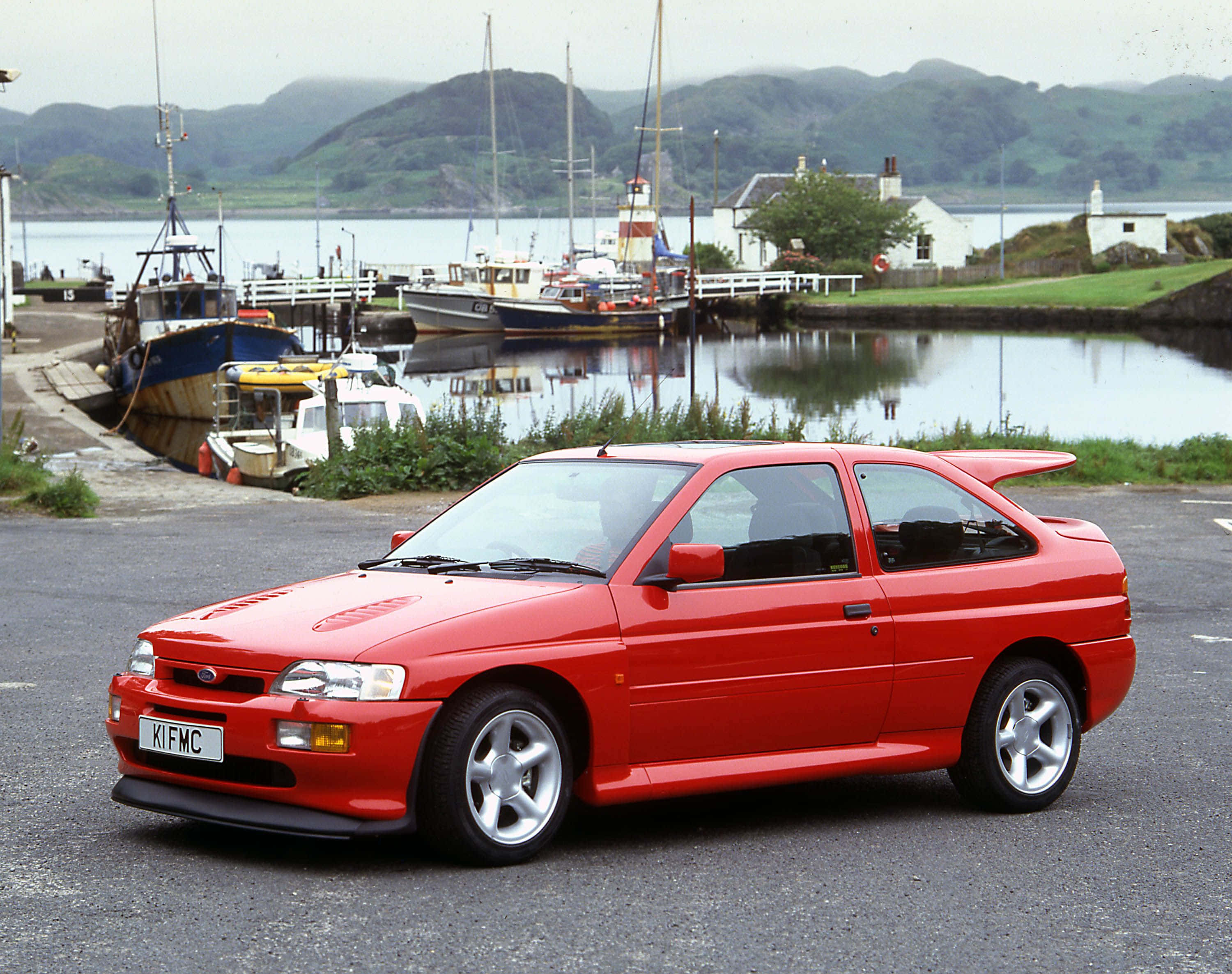 Ford Escort 桌布 壁紙 — 一輛鮮紅色的福特 Escort RS Cosworth 停在柏油路上，旁邊是港灣，背景有船隻和霧氣籠罩的綠色山丘。