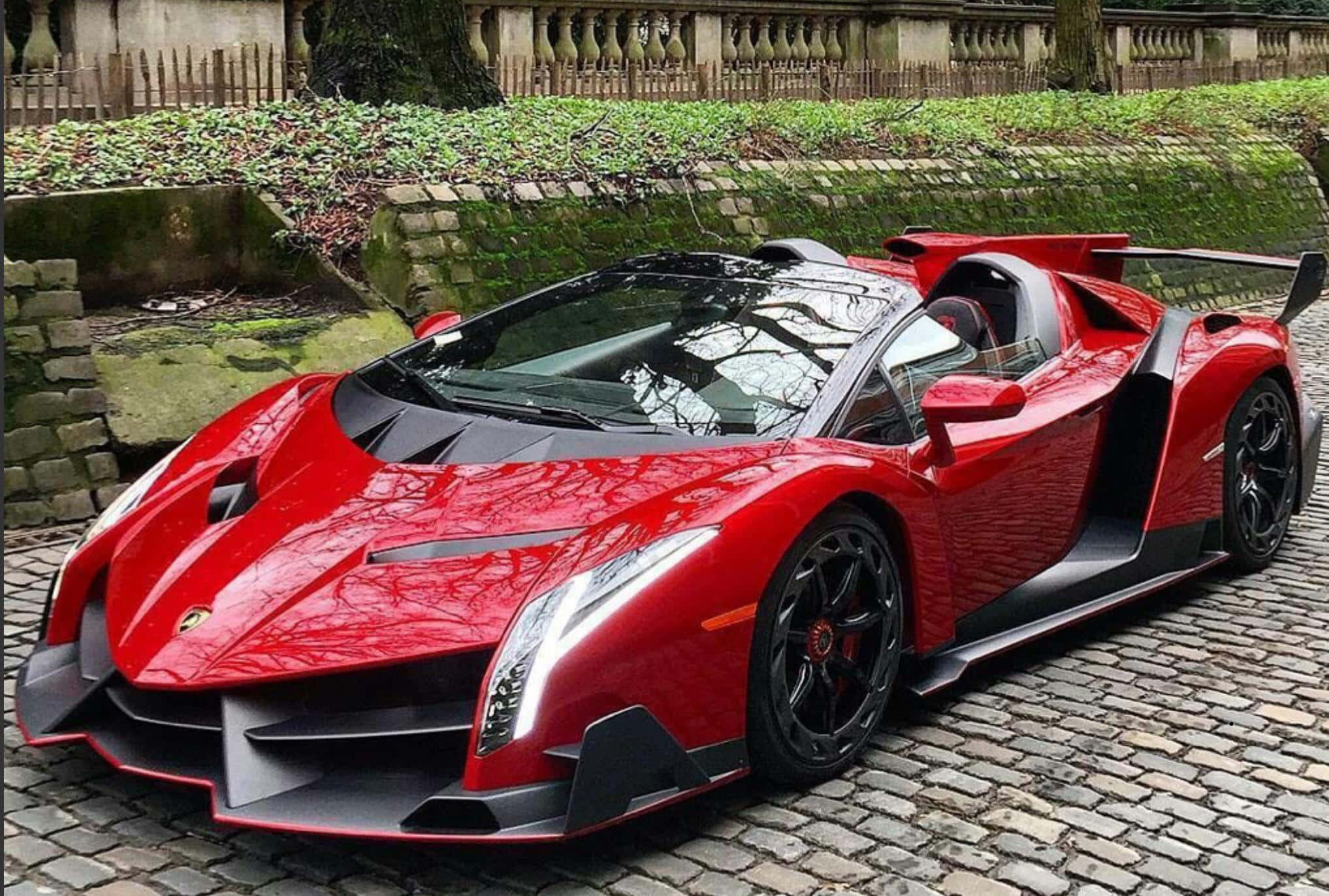Lamborghini Veneno 桌布 壁紙 — 一輛搶眼的紅色藍寶堅尼 Veneno 敞篷跑車停泊在鵝卵石街道上，旁邊是長滿青苔的石牆和綠植。