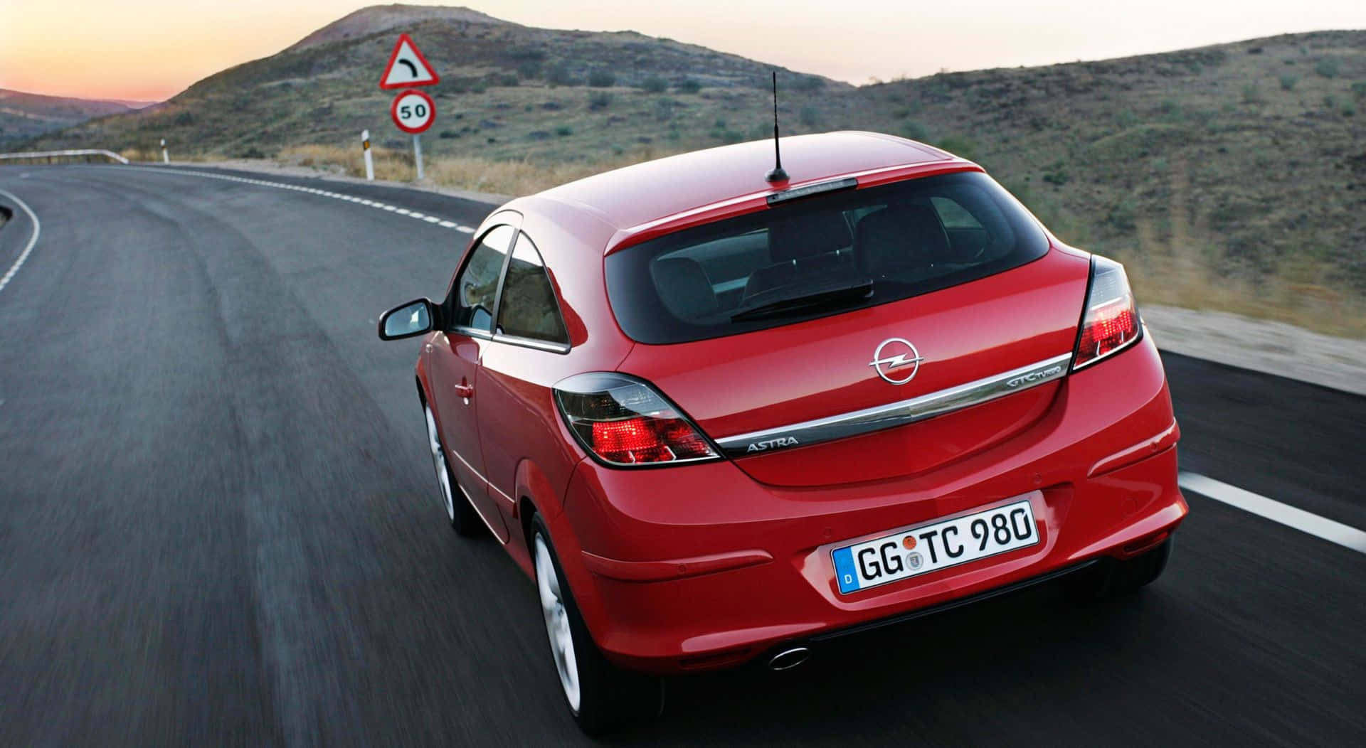 Opel Astra 桌布 壁紙 — 一輛鮮紅色的歐寶雅特雙門跑車在日落時分沿著蜿蜒的山路疾馳，帶有模糊的動態效果和山坡背景。