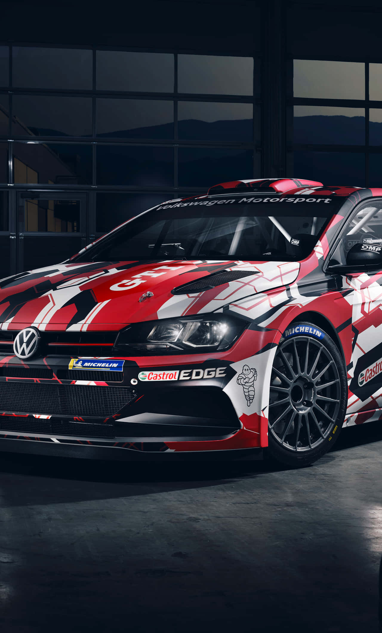 Volkswagen Polo 桌布 壁紙 — 一輛紅白黑色的福斯汽車拉力賽車，帶有贊助商貼紙，停放在光線昏暗的車庫內，窗外可見山脈。
