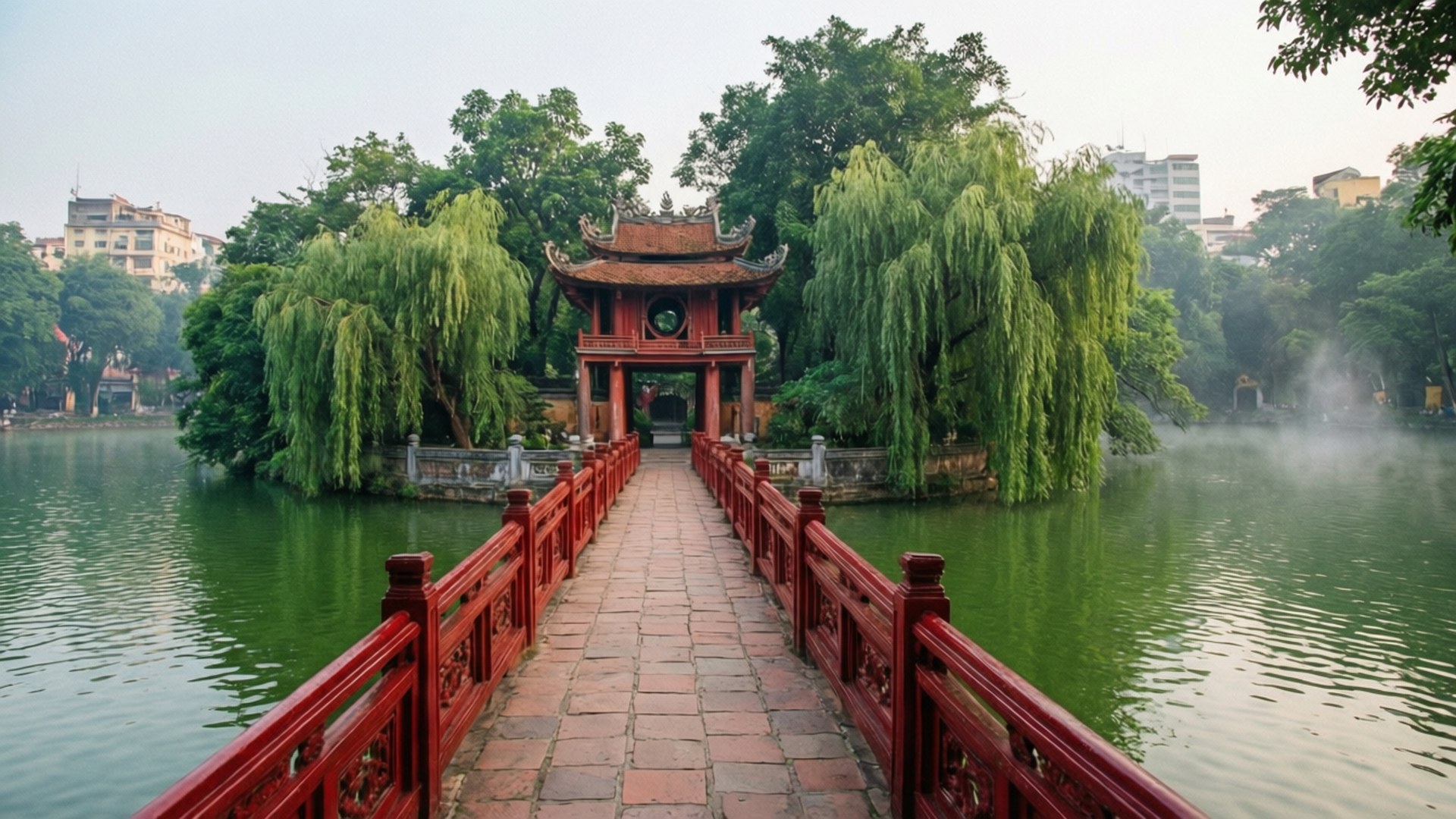 Hanoi Hoan Kiem Lake 壁紙 — 一條紅色木橋通向一個傳統的越南亭閣，周圍環繞著垂柳，位於霧氣瀰漫的綠色湖泊上。