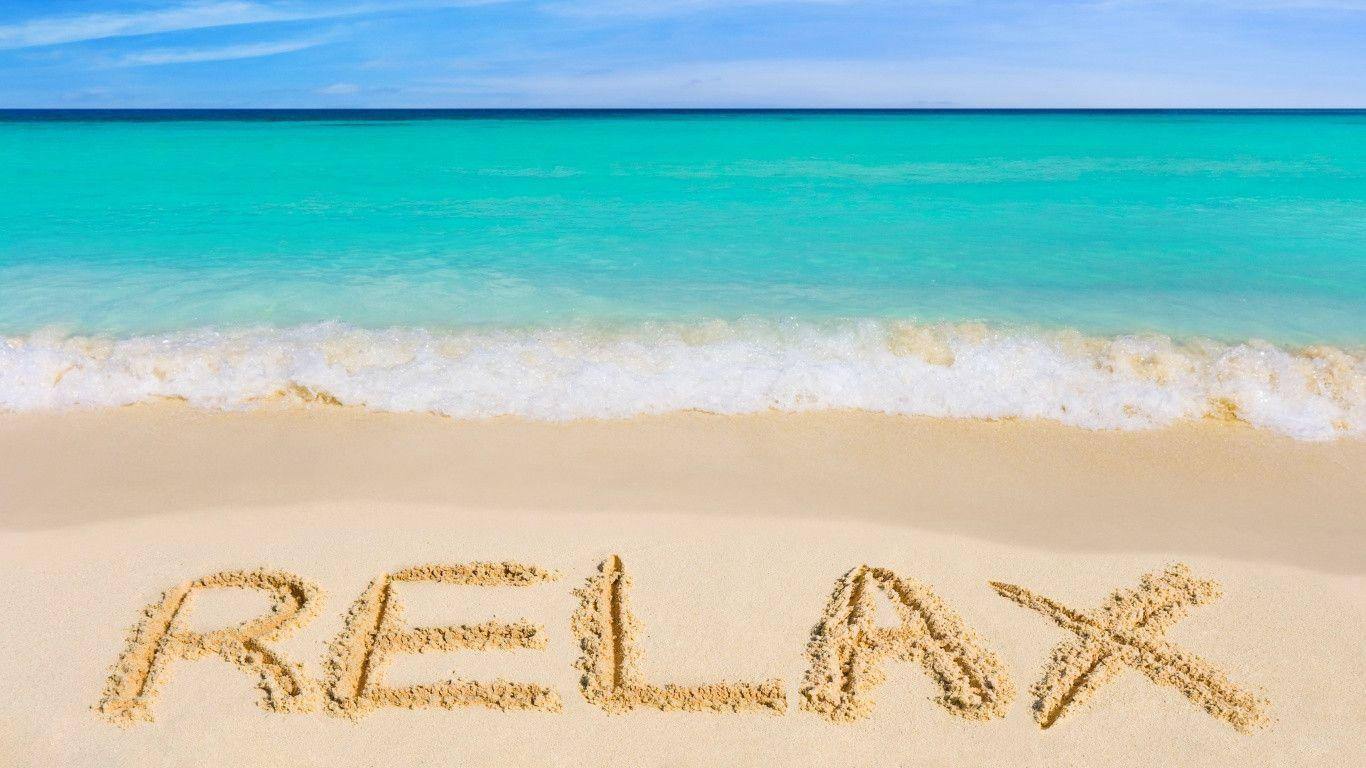 Relaxing 桌布 壁紙 — 「relax」這個字寫在純淨沙灘的沙子上，碧綠色的海浪在清澈藍天下輕輕翻滾。