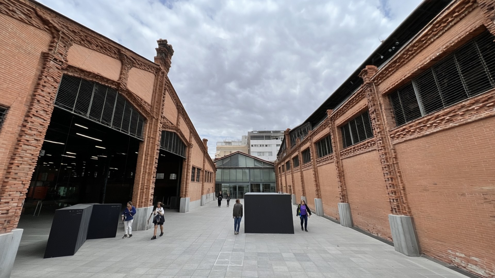 Matadero Art Center 壁紙 — 人們在陰天之下，穿過一個寬敞的庭院，兩旁是高聳的紅磚工業建築。