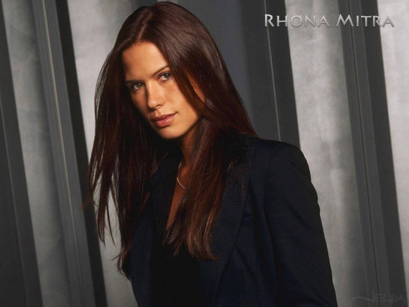 美女 桌布 壁紙 — Rhona Mitra 身穿深色西裝外套，在時尚的灰色垂直條紋背景前自信地擺姿勢，搭配柔和的攝影棚燈光。