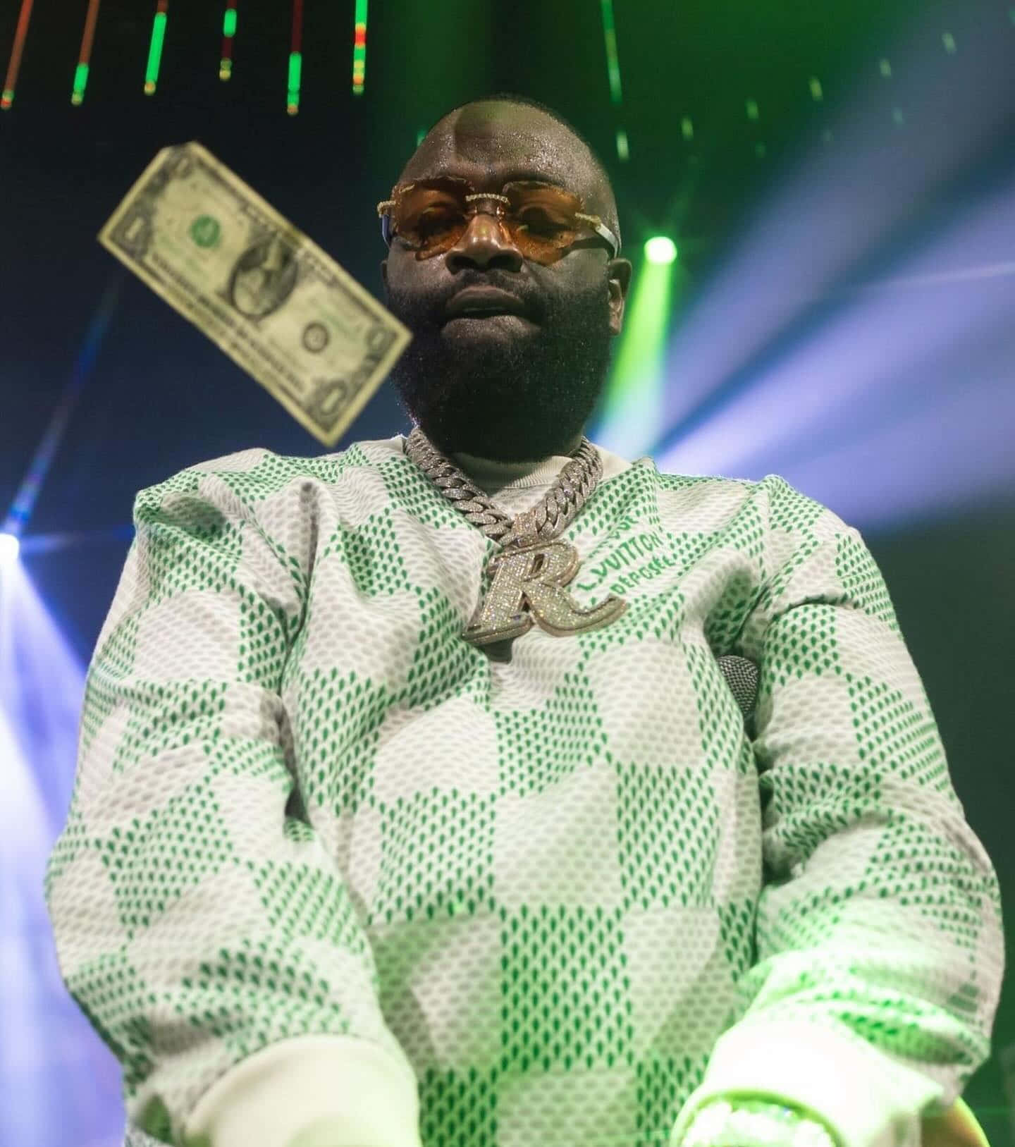  壁紙 — Rick Ross 自信地站在舞台上，身穿綠色和白色圖案服裝，頭頂漂浮著一張美元鈔票，在色彩繽紛的舞台燈光下。
