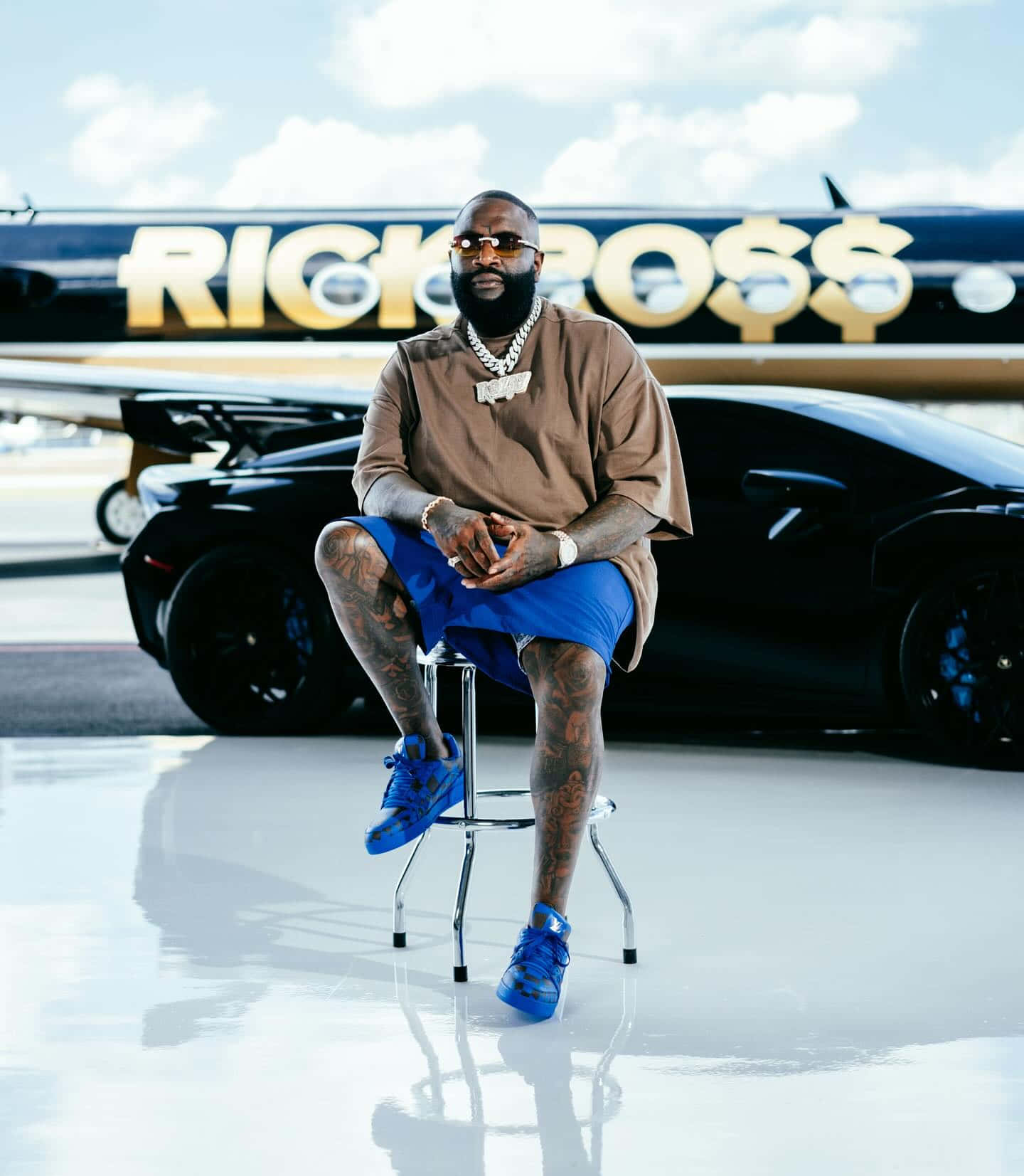  壁紙 — Rick Ross 自信地坐在凳子上，身後是他的私人飛機和豪華汽車，天空晴朗。