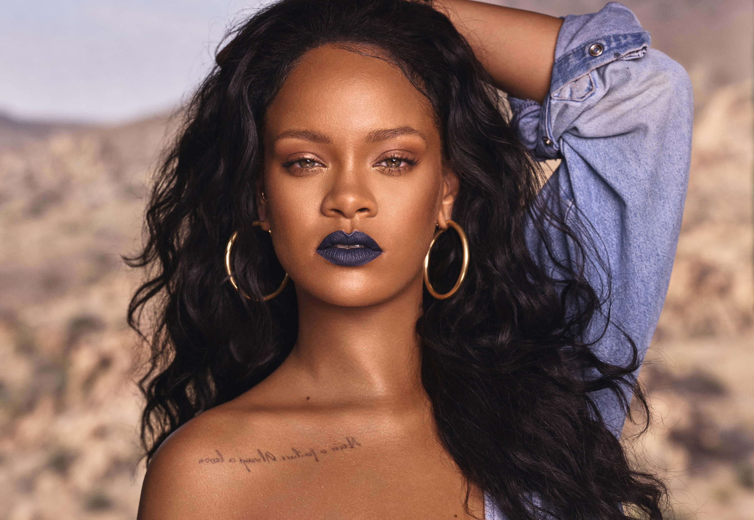 蕾哈娜 桌布 壁紙 — Rihanna 在沙漠中自信地擺姿勢，塗著深藍色唇膏，戴著金色耳環，背景是岩石。