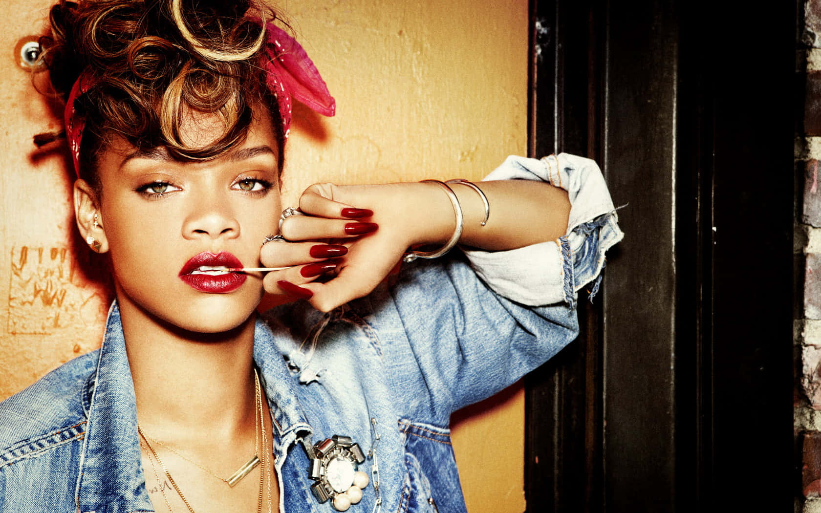 蕾哈娜 桌布 壁紙 — Rihanna 在溫暖燈光下，以紋理牆壁為背景，擺出大膽紅唇和牛仔外套的姿勢。