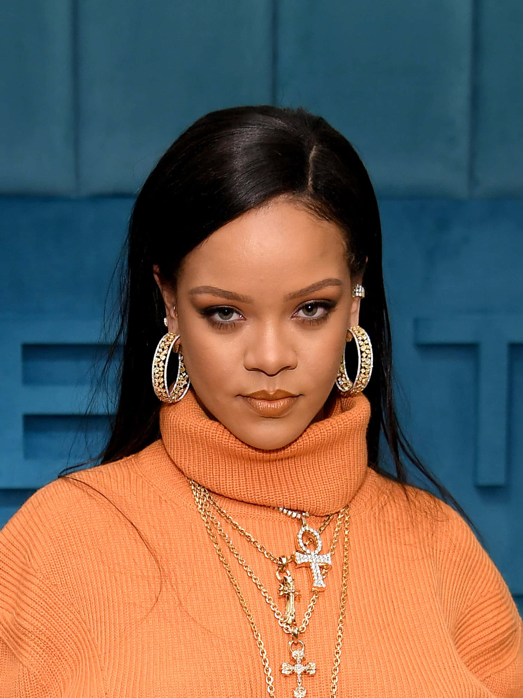 Celebrity 桌布 壁紙 — Rihanna 自信地凝視著，身穿橙色高領毛衣，搭配金色項鍊和大圈耳環，背景是藍色紋理牆。
