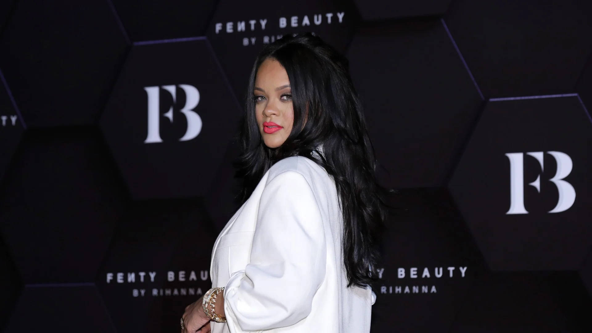 Fenty 桌布 壁紙 — Rihanna 自信地擺姿勢，身穿白色西裝外套，背景是黑色的六角形 Fenty Beauty 背景。