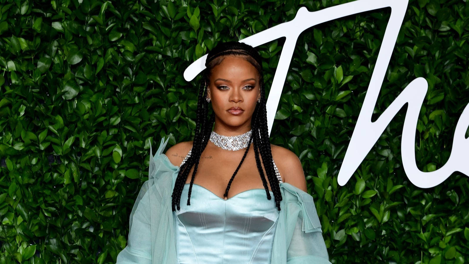 Fenty 桌布 壁紙 — Rihanna 自信地擺姿勢，身穿淺藍色緞面長袍，背景是茂密的綠色樹籬牆，上面有白色字體。