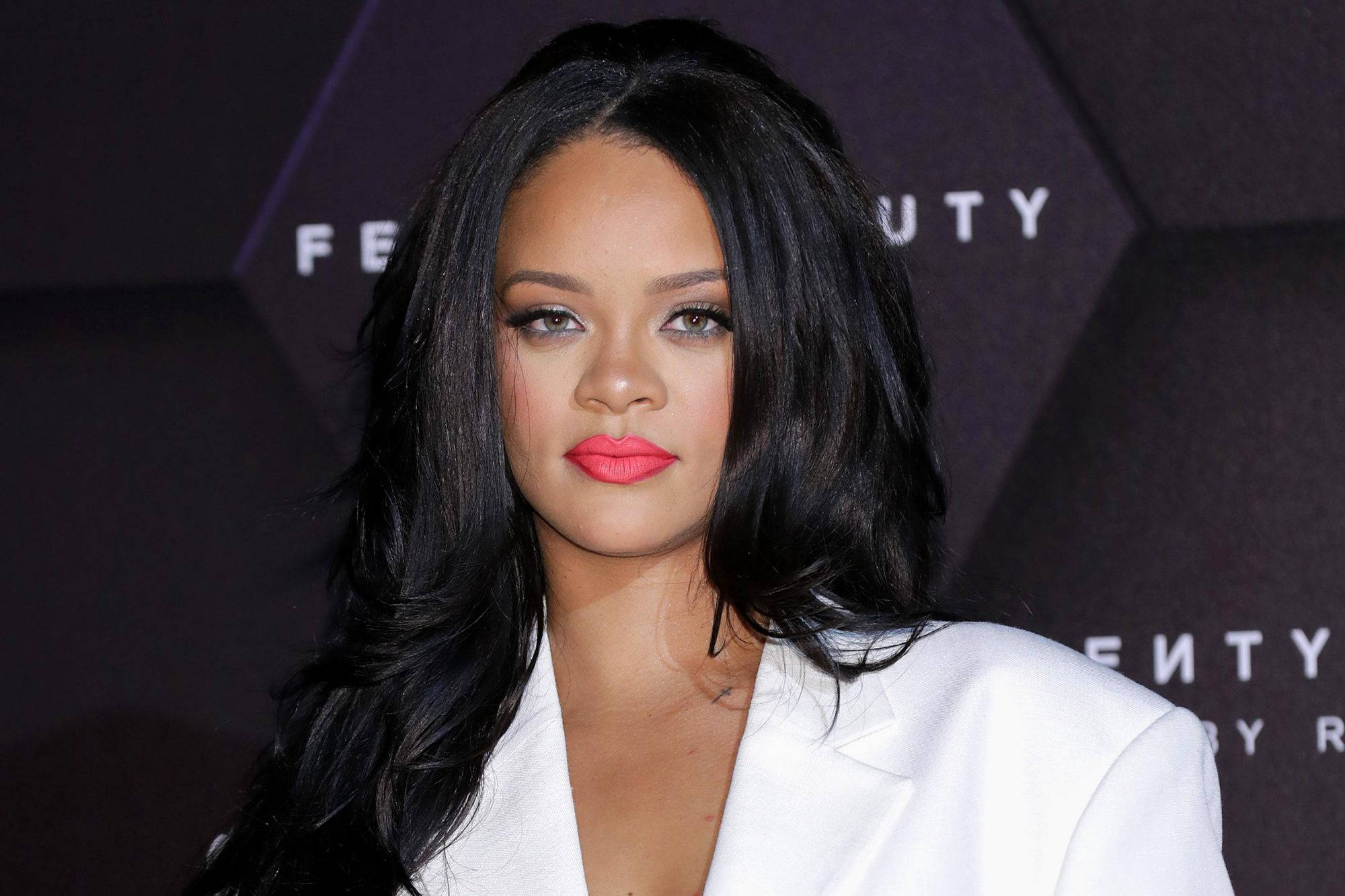 蕾哈娜 桌布 壁紙 — Rihanna 自信地穿著白色西裝外套，在深色品牌背景前擺姿勢，擁有鮮豔的粉紅嘴唇和豐盈的黑髮。