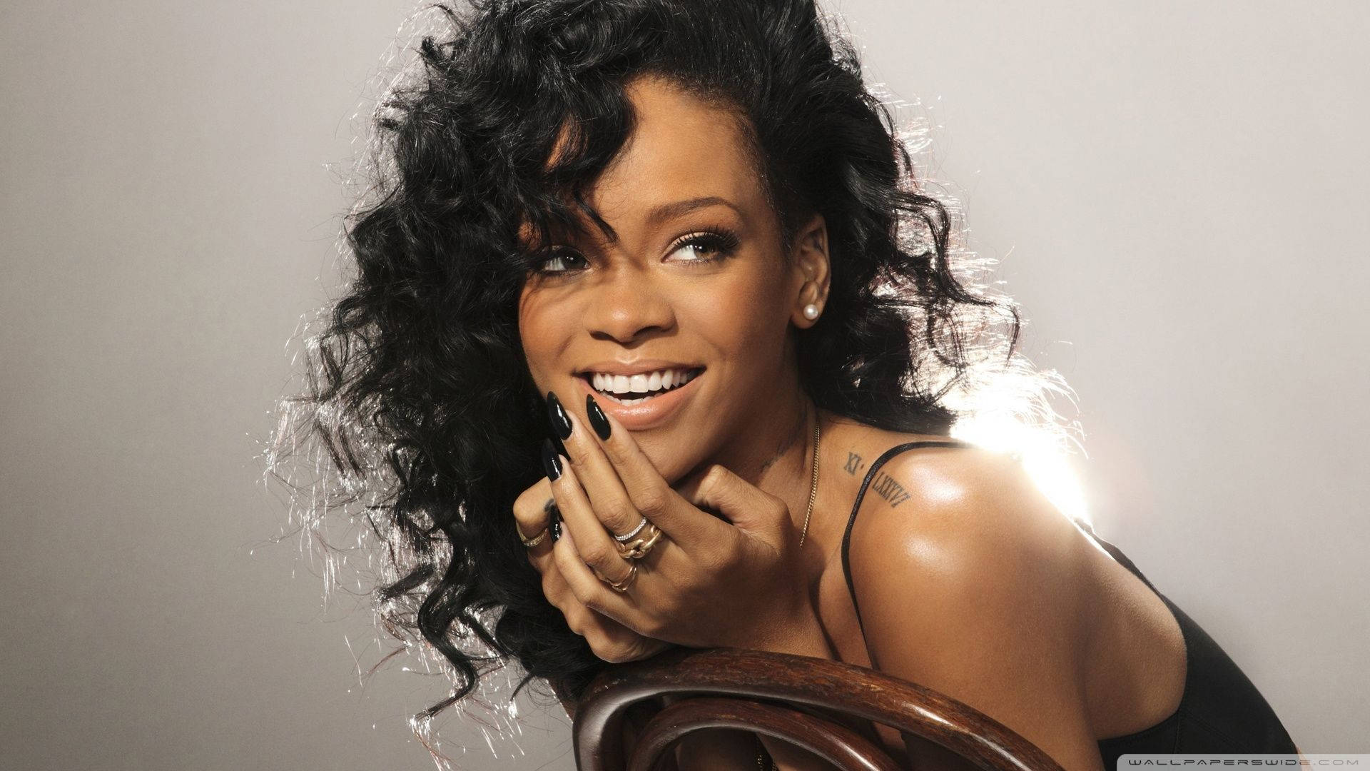 蕾哈娜 桌布 壁紙 — Rihanna 露出燦爛笑容，黑色捲髮和綠色指甲，背景為柔和灰色。