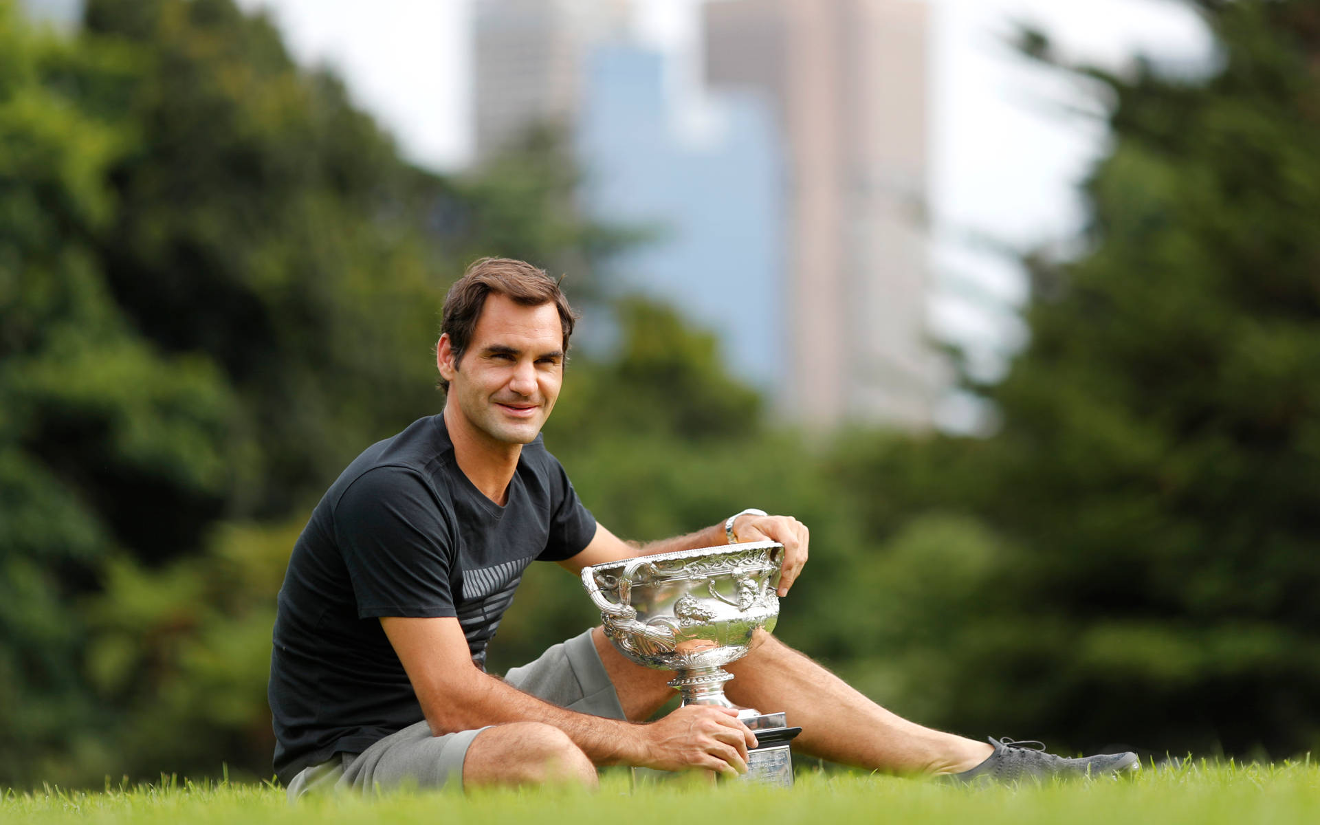 roger 桌布 壁紙 — Roger Federer 坐在草地上，手持銀色獎盃，背後的城市摩天大樓模糊不清。