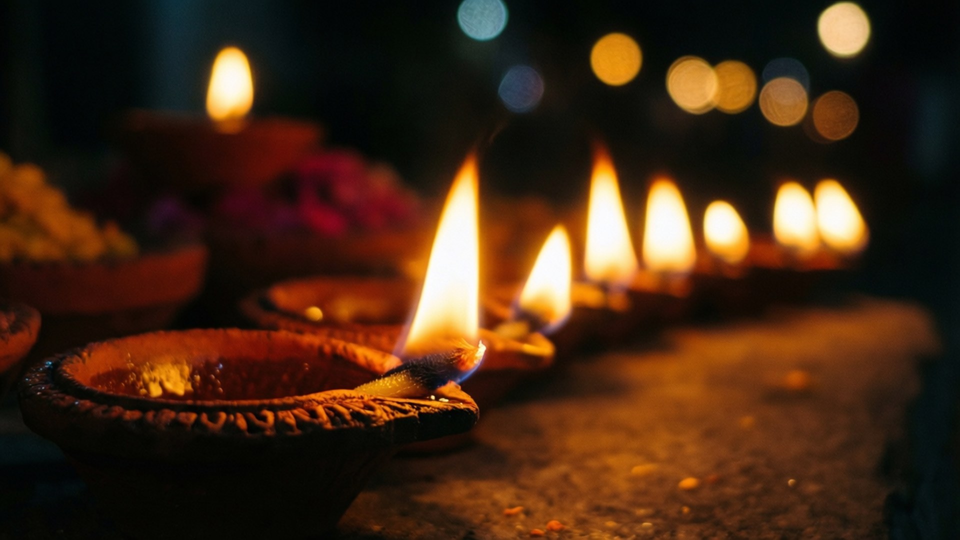 Glowing Diyas at Night 壁紙 — 一排赤陶迪亞，閃爍的火焰在黑暗模糊的背景中投射出溫暖的金色光芒，帶有散景燈光。