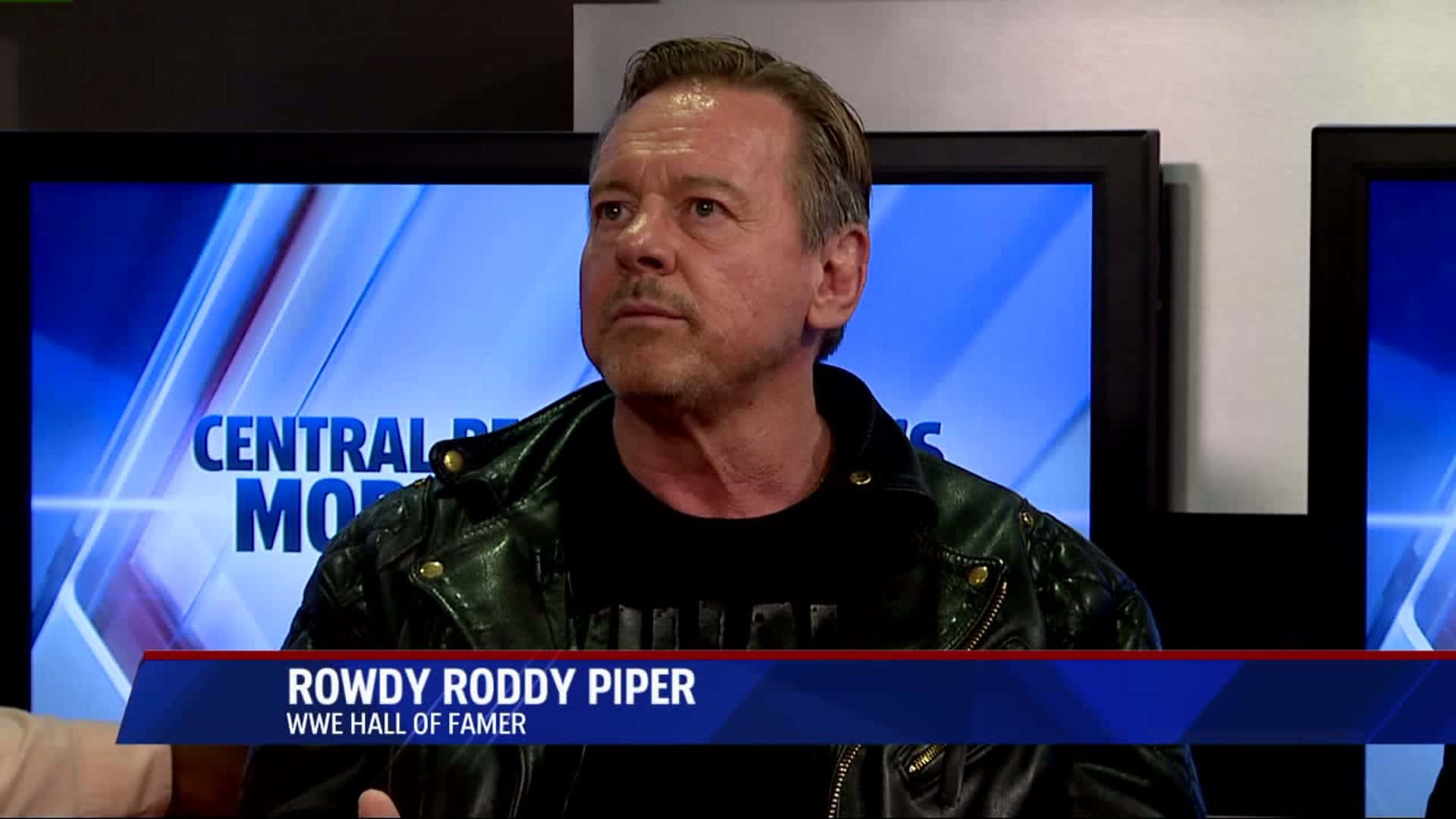 摔角 桌布 壁紙 — Rowdy Roddy Piper 身穿黑色皮夾克，坐在顯示 'Central News' 的藍色電視螢幕前，表情嚴肅。