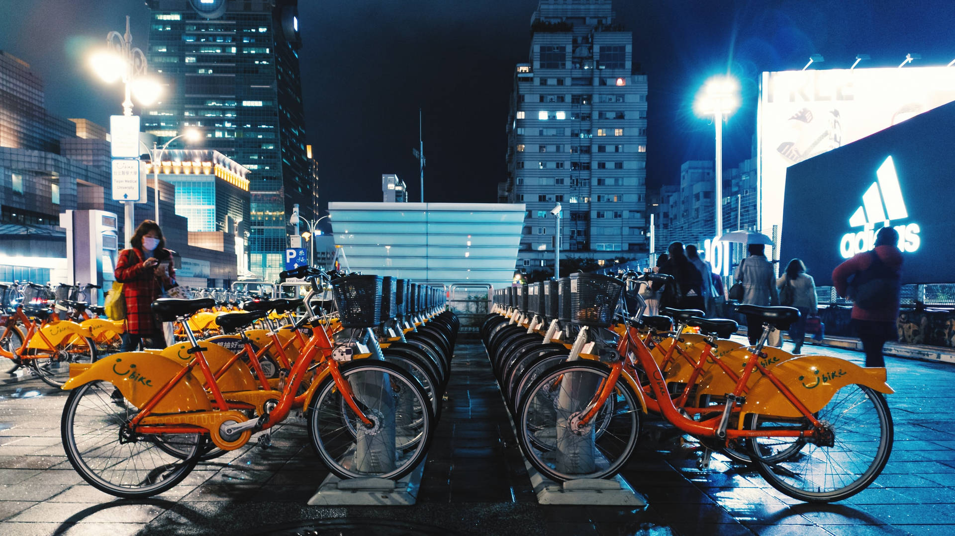 一排橙色共享單車停在濕漉漉的城市街道上，夜晚有發光的招牌和行人。 — 台北 壁紙