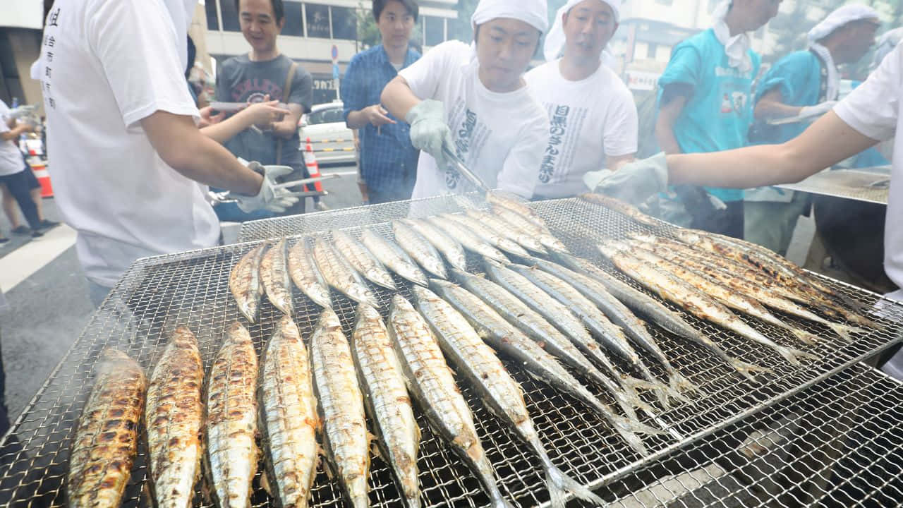  壁紙 — 一排排烤魚在大型街頭烤架上滋滋作響，身穿白襯衫和手套的廚師在旁觀者圍繞下照料著煙燻美食。