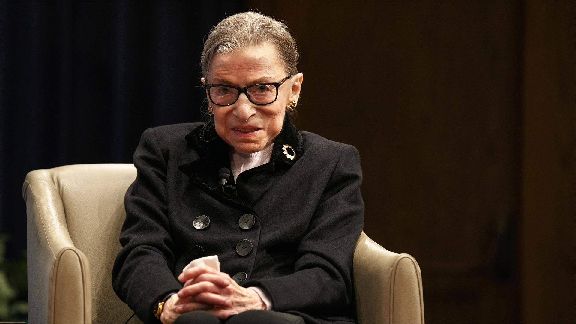 露絲·拜德·金斯伯格 桌布 壁紙 — Ruth Bader Ginsburg 坐在米色扶手椅上，穿著黑色外套和眼鏡，表情嚴肅。