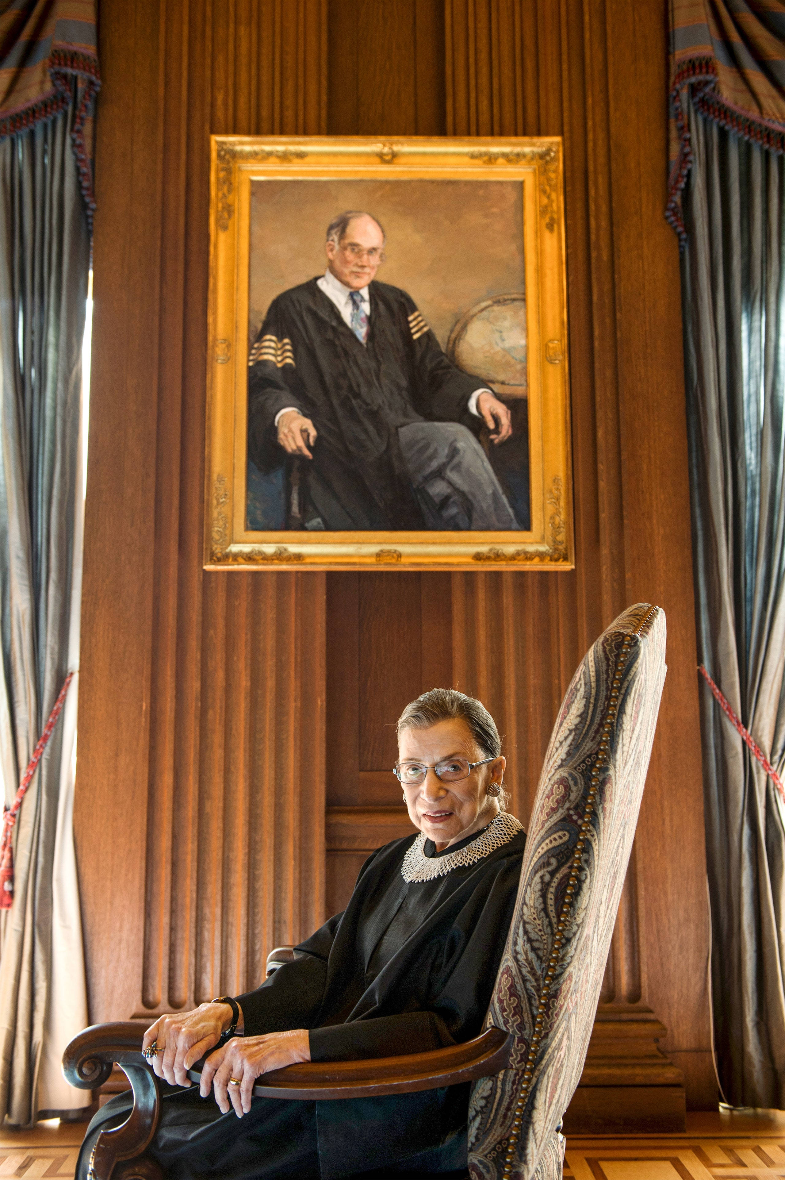 Ruth Bader Ginsburg 桌布 壁紙 — Ruth Bader Ginsburg 坐在木椅上，下方掛著她身穿司法袍的肖像，背景是華麗的木鑲板和垂簾。