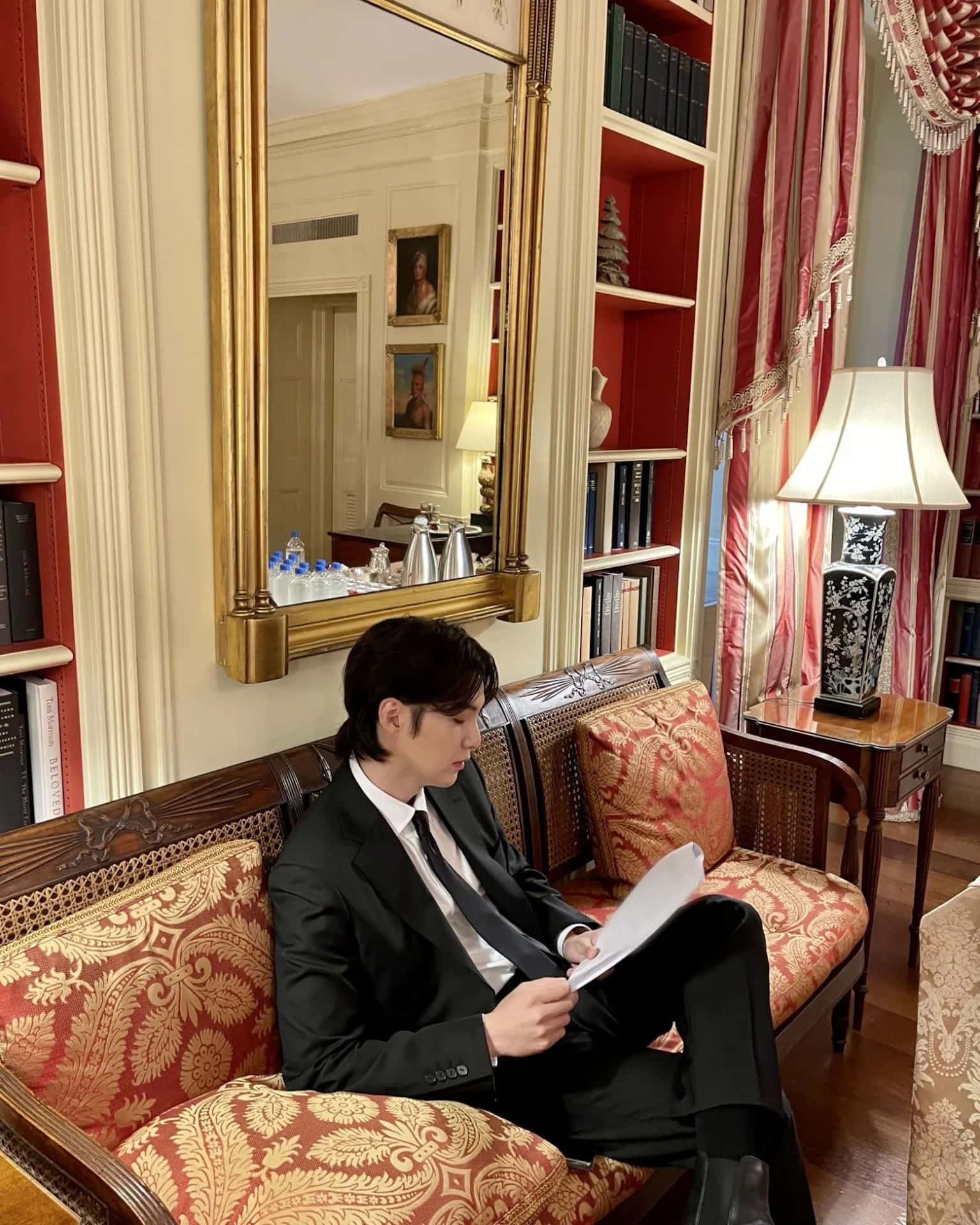  壁紙 — 一名身穿黑色西裝的男子坐在一張華麗的紅金圖案沙發上，在一個裝飾華麗、有書架和大鏡子的房間裡閱讀文件。