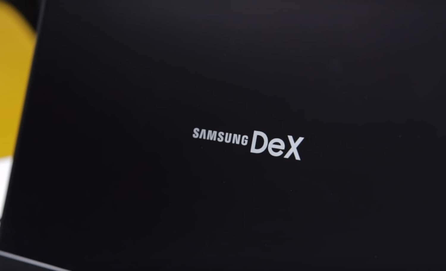 三星 桌布 壁紙 — Samsung DeX 標誌的特寫，白色文字印在光滑的黑色表面上，帶有黃色背景元素。