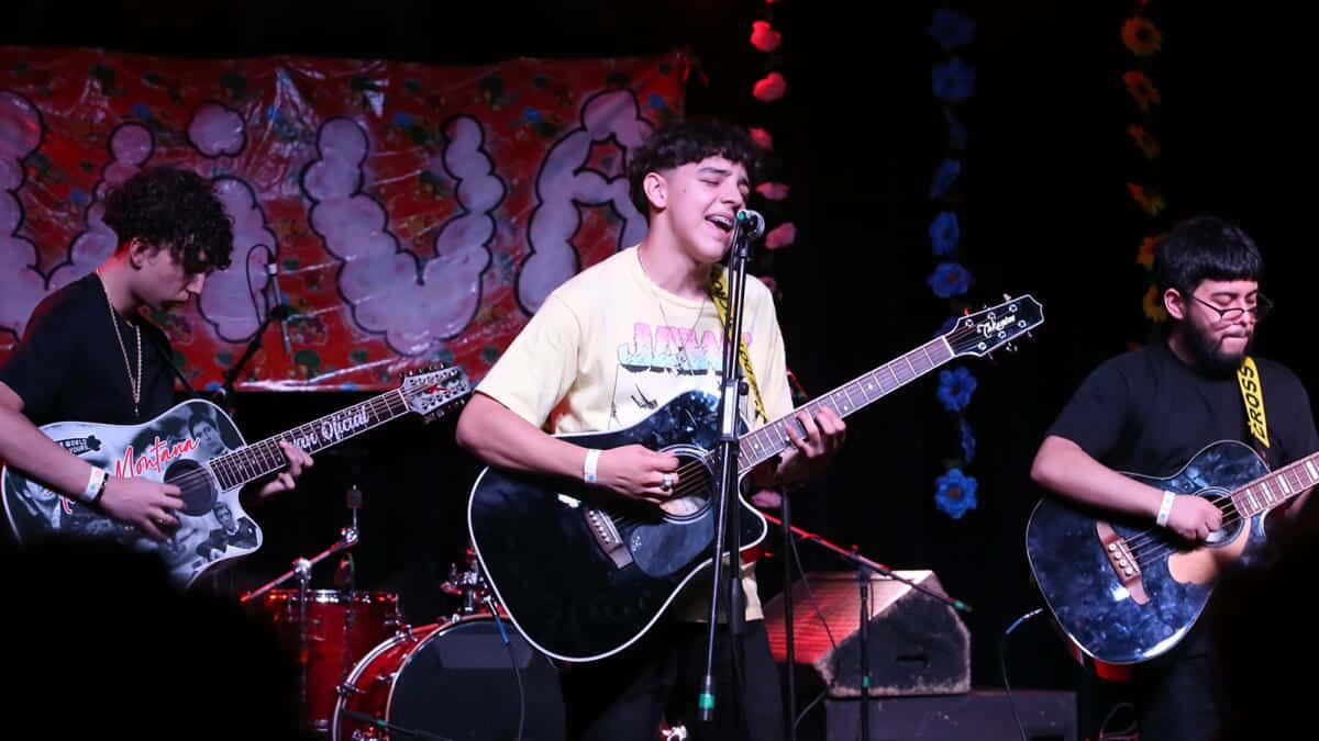  壁紙 — 三位年輕音樂家在色彩繽紛的背景燈光下，帶著吉他在舞台上熱情演出。