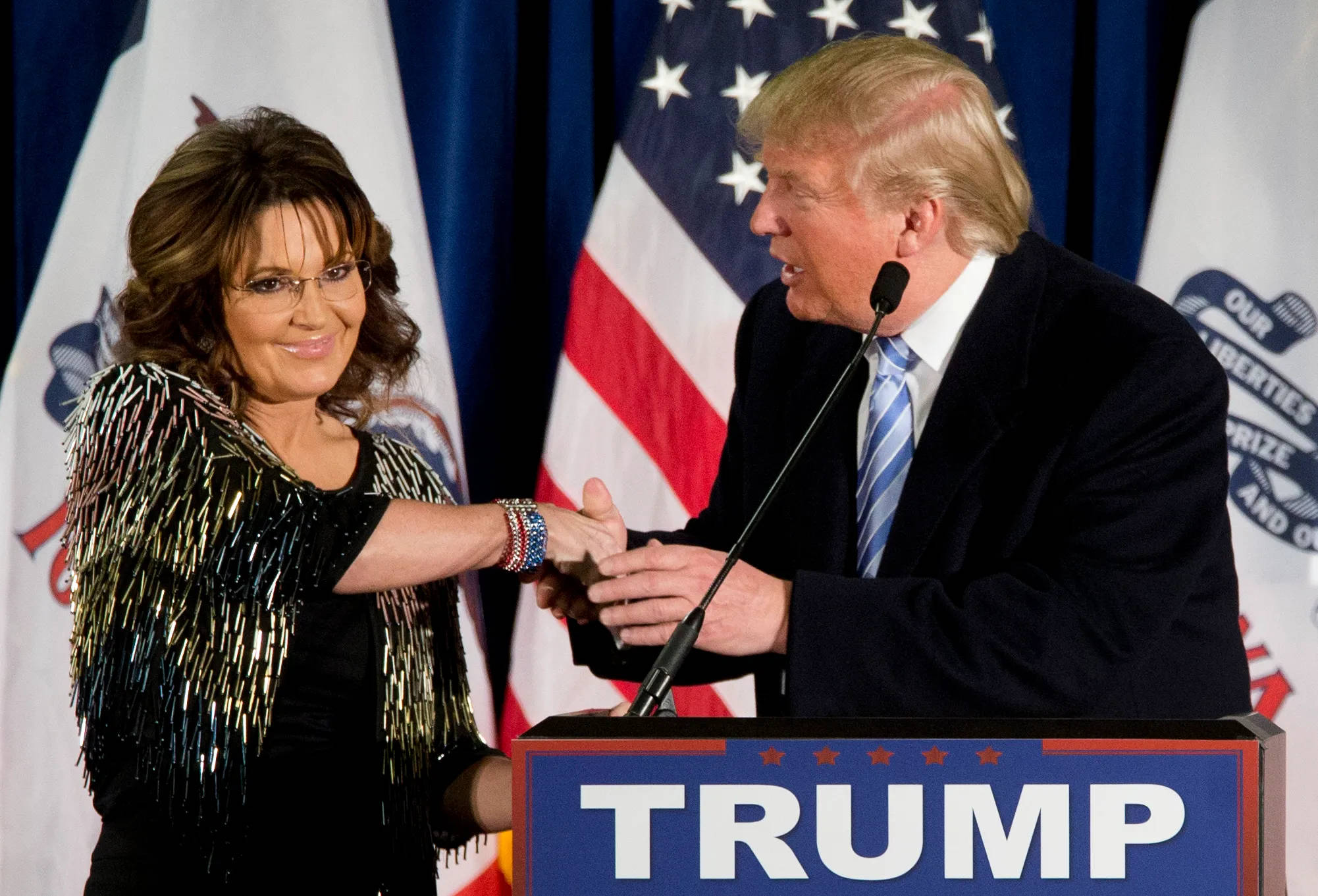 川普 桌布 壁紙 — Sara Lee 和 Donald Trump 在競選活動中握手，背後有美國國旗和 Trump 講台。