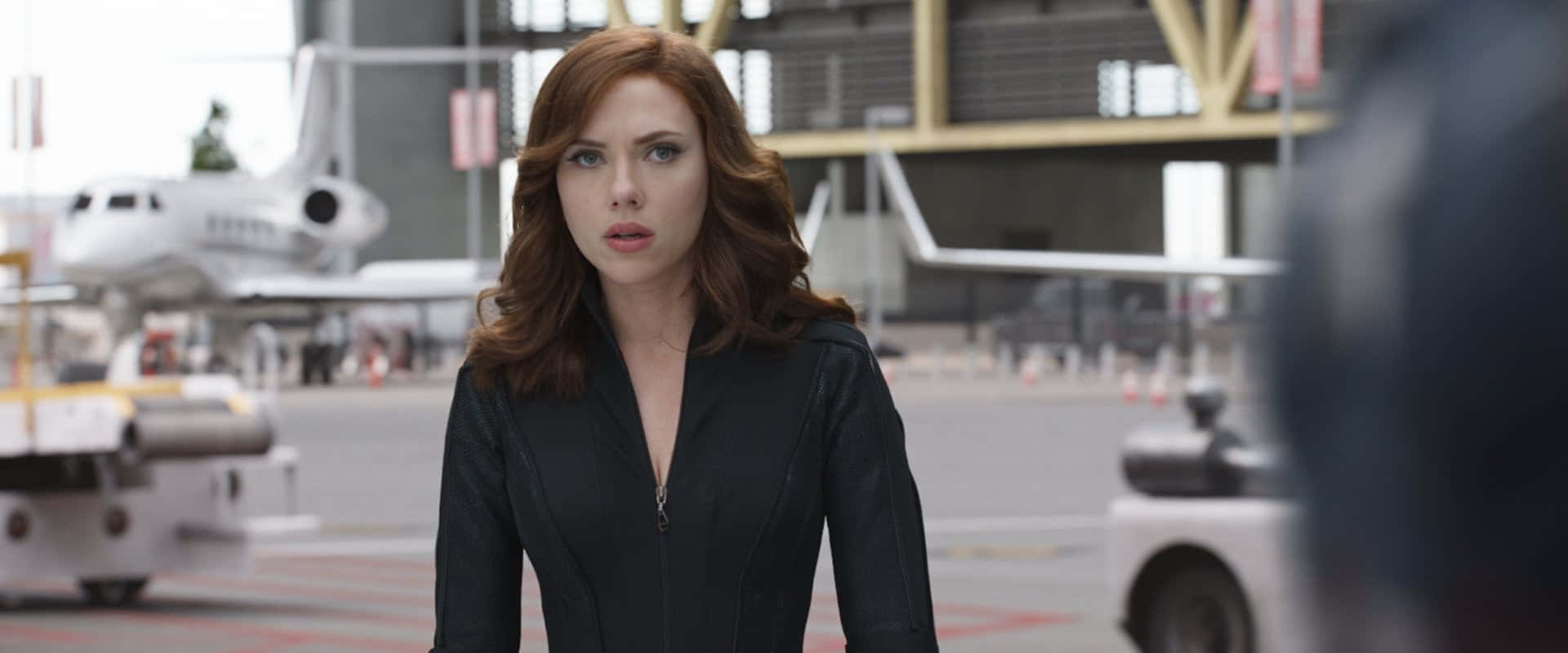 Black Widow 桌布 壁紙 — Scarlet Witch 站在機場停機坪，身穿黑色外套，背景有私人飛機和地面車輛。
