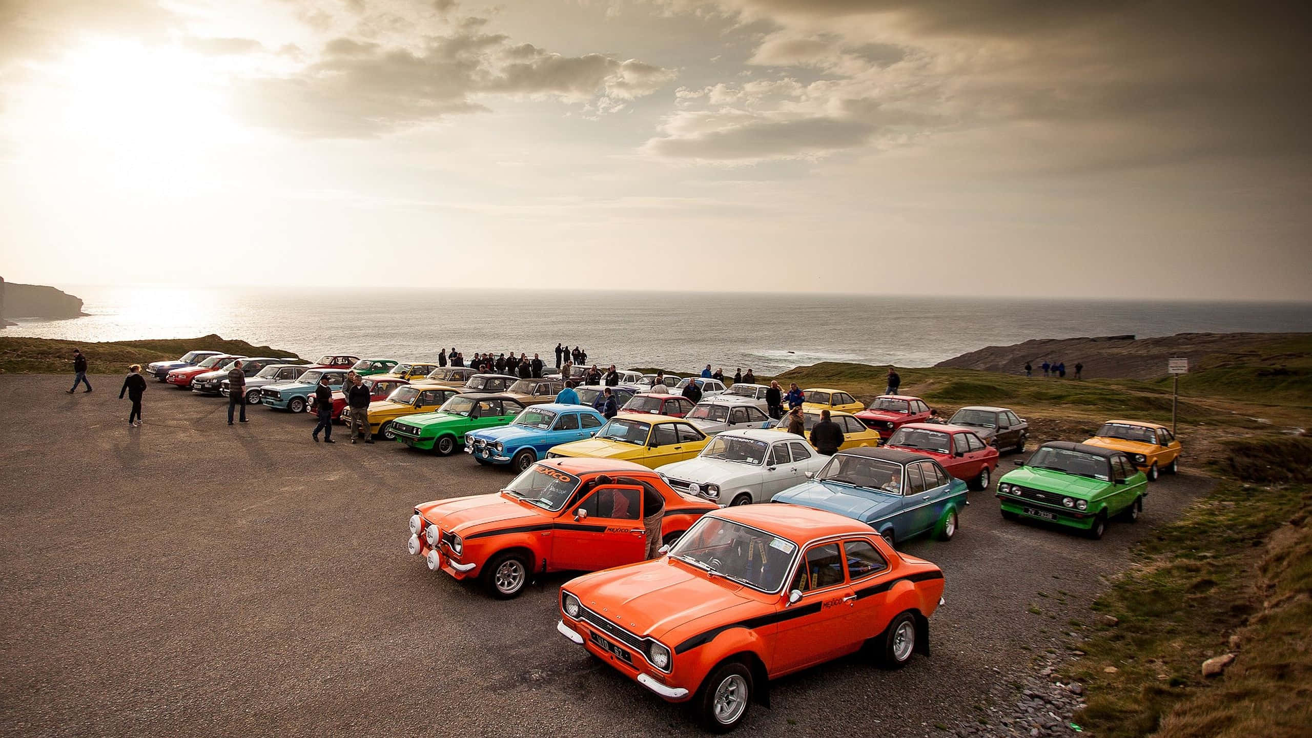 Ford Escort 桌布 壁紙 — 色彩繽紛的經典汽車停泊在俯瞰日落時分海洋的海岸懸崖上，人們正在欣賞它們。