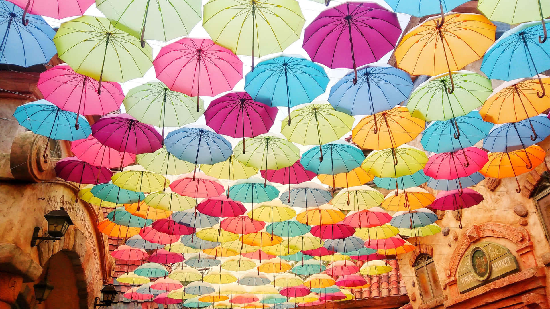  壁紙 — 色彩繽紛的雨傘在陽光普照的歐式街道上形成節日般的天篷。