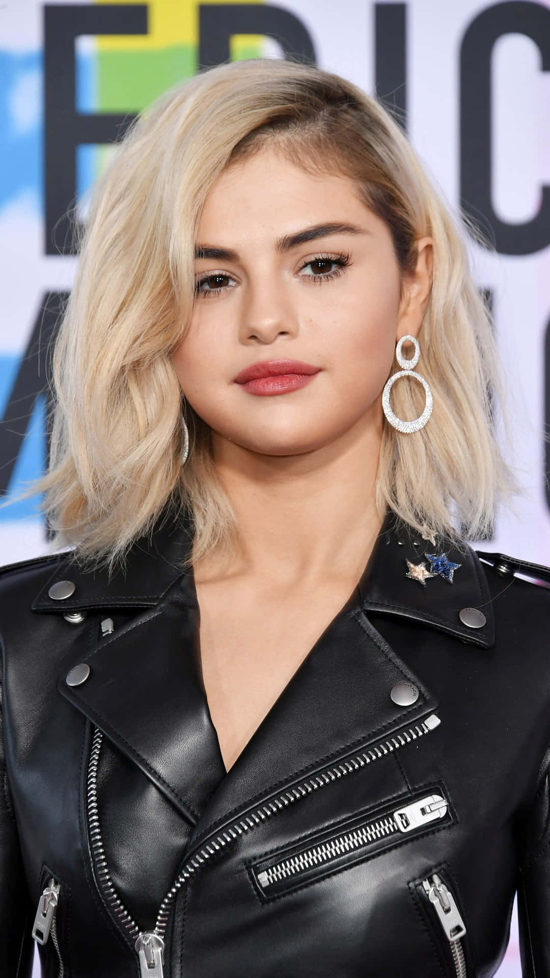 桌布 壁紙 — Selena Gomez 在活動中穿著帶有星星補丁的黑色皮夾克和大圈耳環擺姿勢。