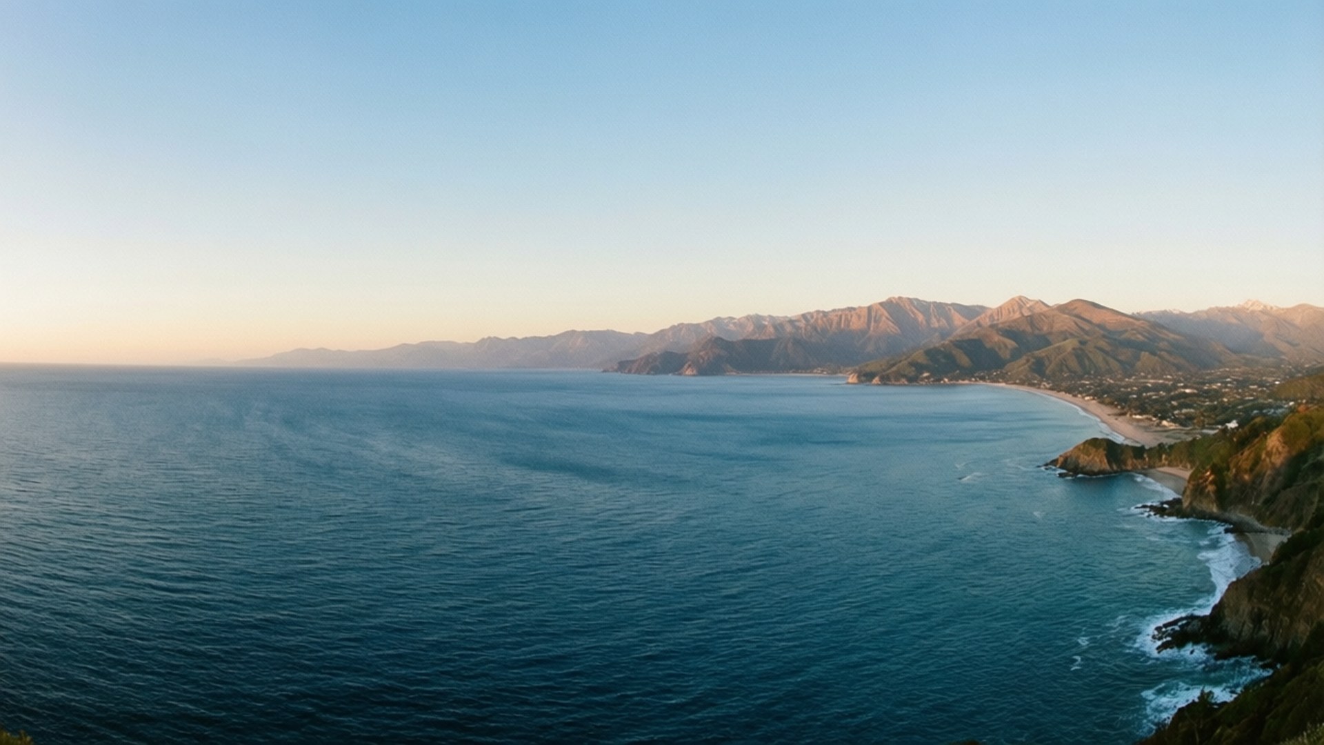 Panoramic Coastline Mountains 壁紙 — 寧靜的海岸景色，深藍色的海洋與金色的沙灘相遇，崎嶇的懸崖和遠處的山脈在晴朗的天空下。