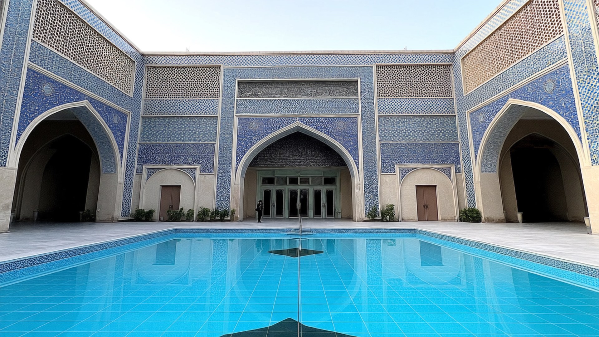 Al-Rahman Mosque Detail 壁紙 — 一個寧靜的庭院設有一個明亮的藍色瓷磚泳池，反射出帶有拱門的複雜藍色和米色伊斯蘭建築。